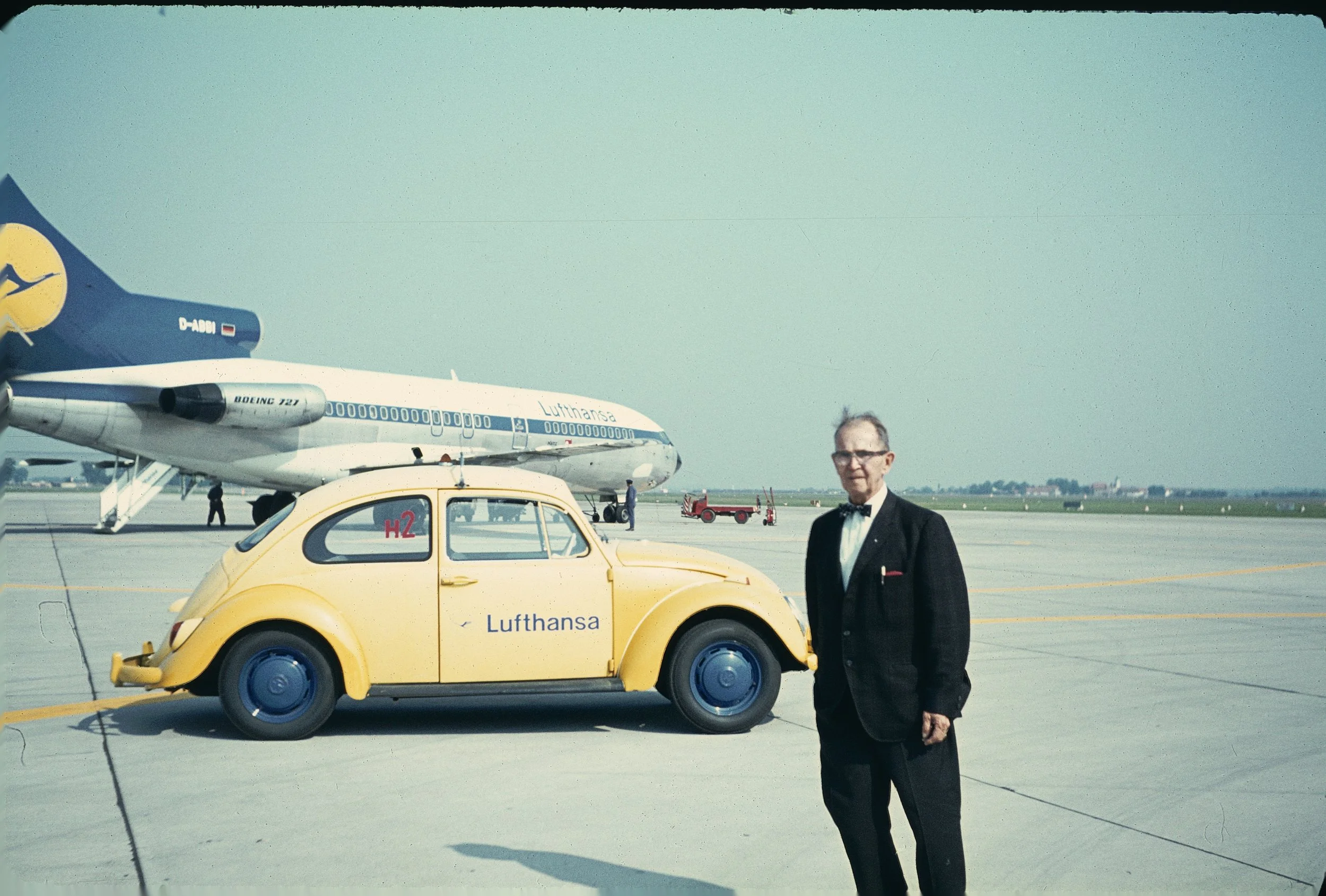 Man in a tuxedo standing next to a vintage yellow Lufthansa car on an airport tarmac with a Lufthansa Boeing 747 airplane in the background.