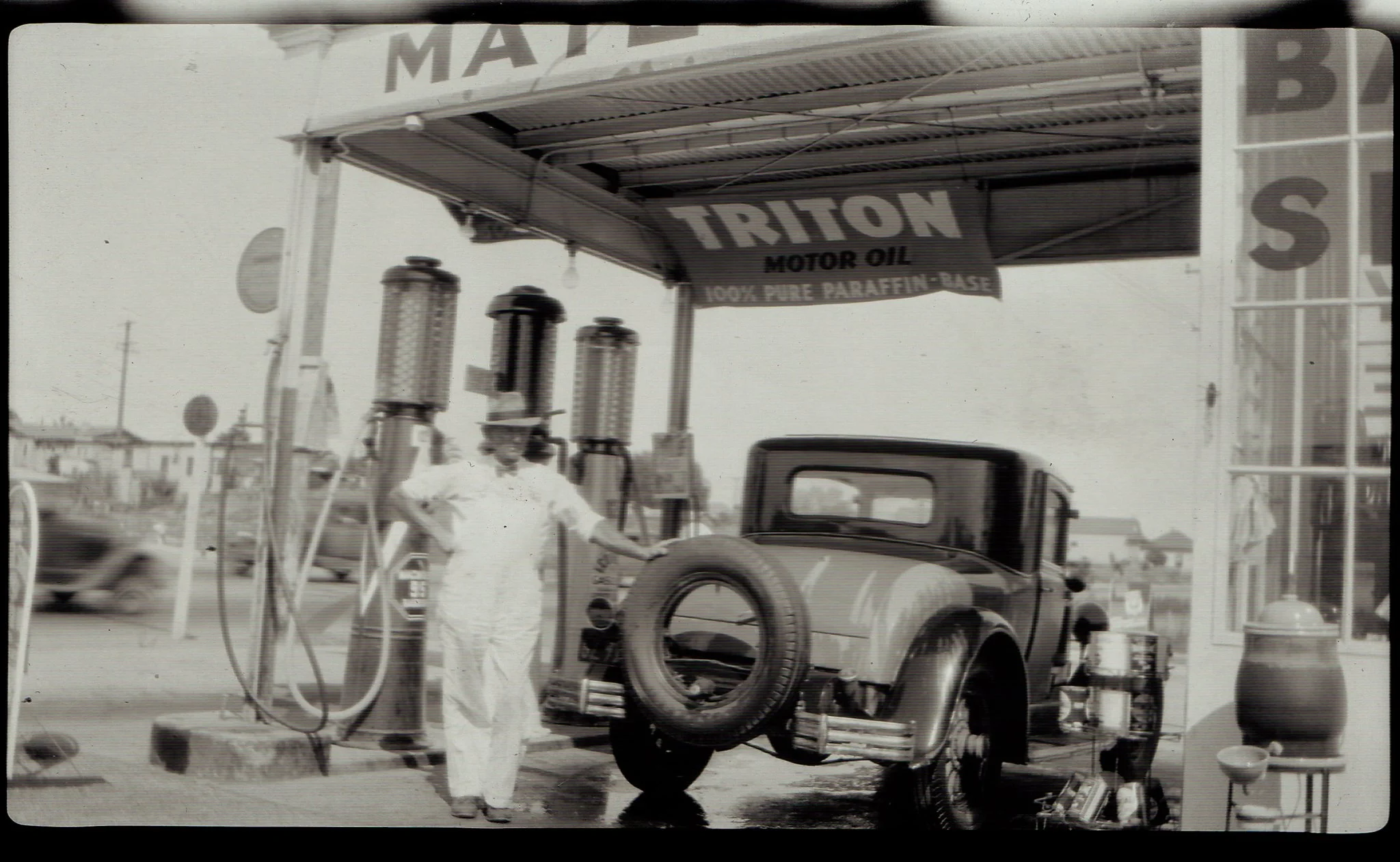 A vintage black and white photo of an outdoor gas station with a car being fueled. There are fuel pumps and a sign for motor oil in the background, with a man in white attire standing near the pumps.
