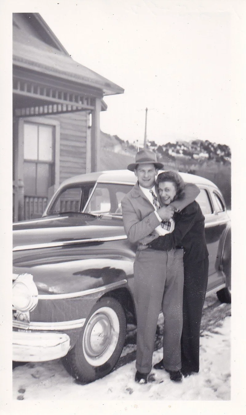 A black-and-white photo of a young man and woman embracing in front of a vintage car on a snowy ground, with a house and hills in the background.