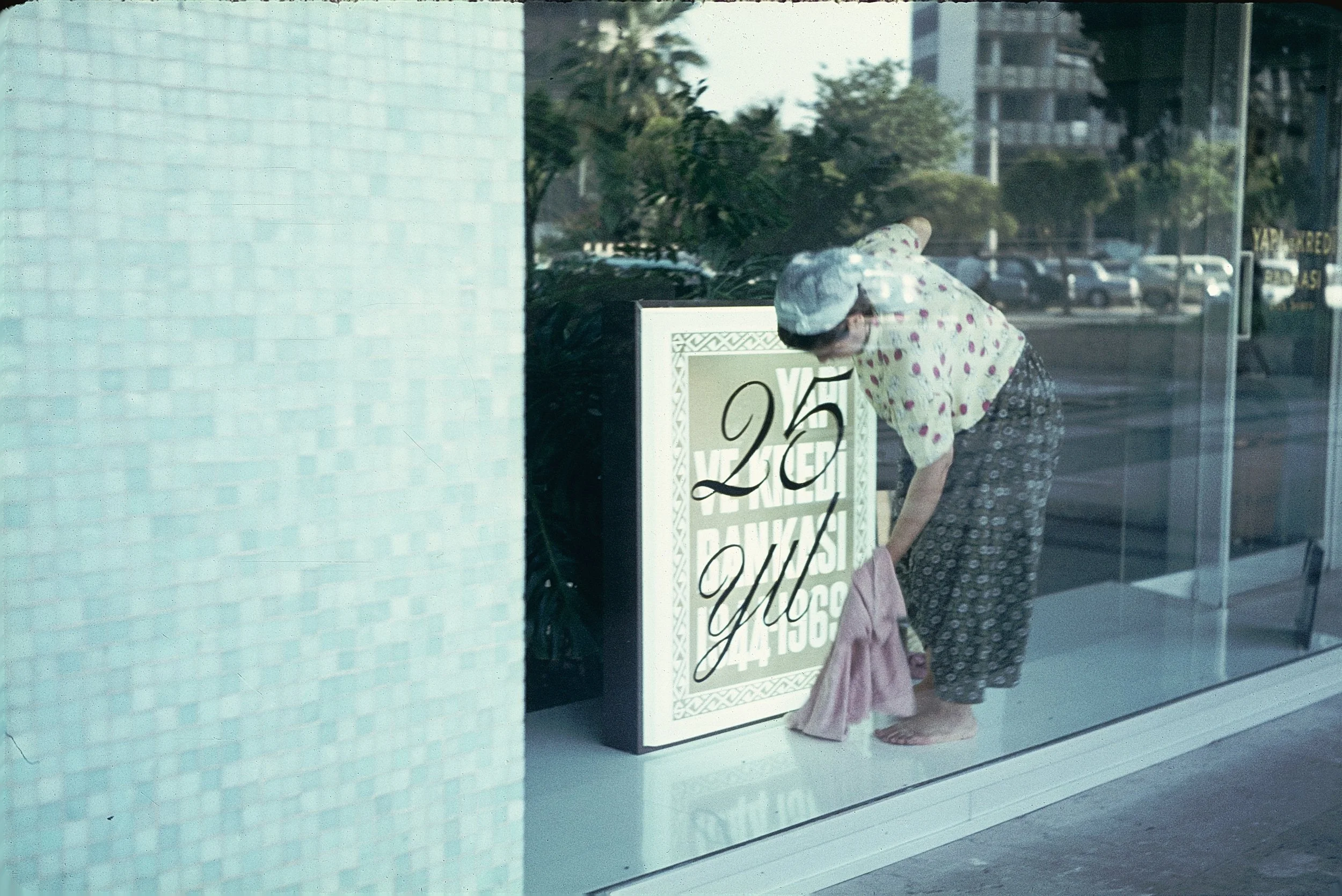 A woman dressed in a vintage polka dot dress and a towel wrapped around her head is hanging laundry on a clothesline outside a glass storefront, with cars and trees visible in the background.
