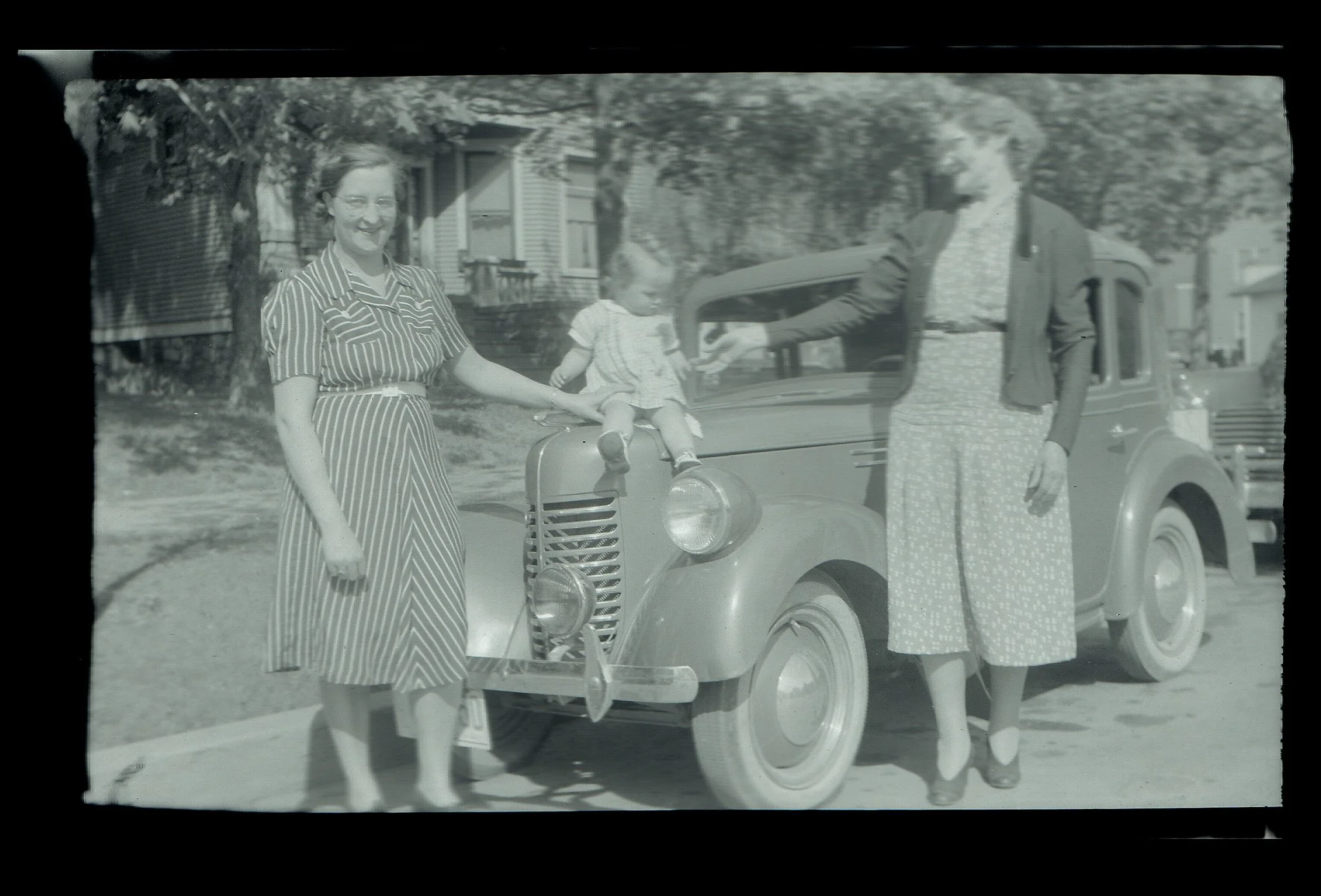 Black and white photo of a woman in a striped dress holding hands with a young girl sitting on a vintage car, with another woman standing nearby, in a residential neighborhood with trees and houses in the background.