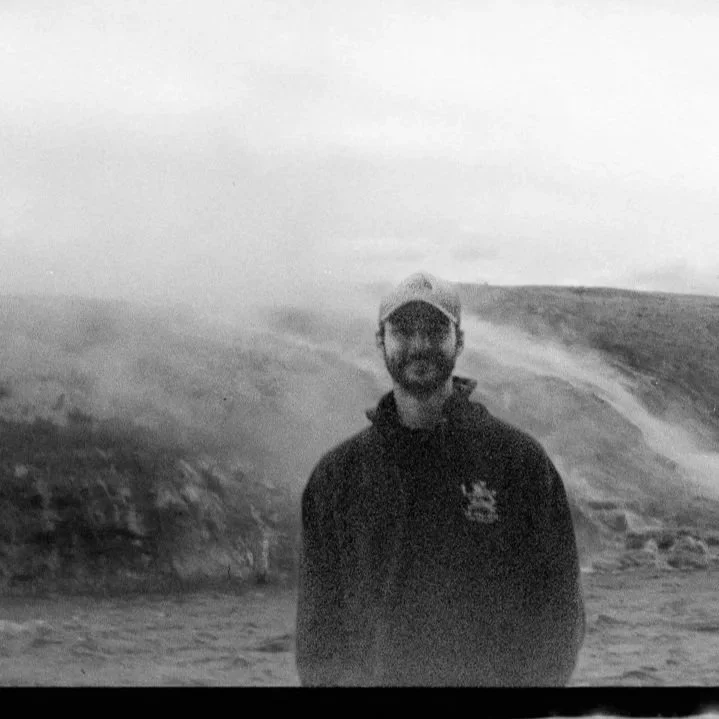 Man standing outdoors on a rocky terrain with mountains and cloudy sky in the background.