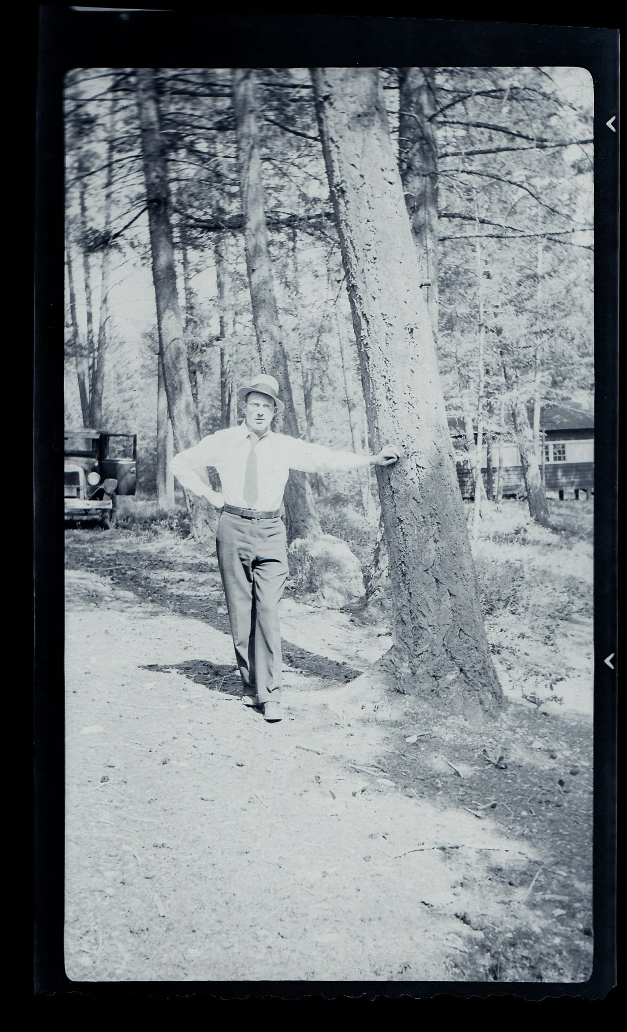 Black and white photograph of a man standing outdoors, leaning on a large tree with his right arm, wearing a light-colored shirt, dark trousers, and a hat. There are trees and wooden houses and a vintage vehicle in the background.