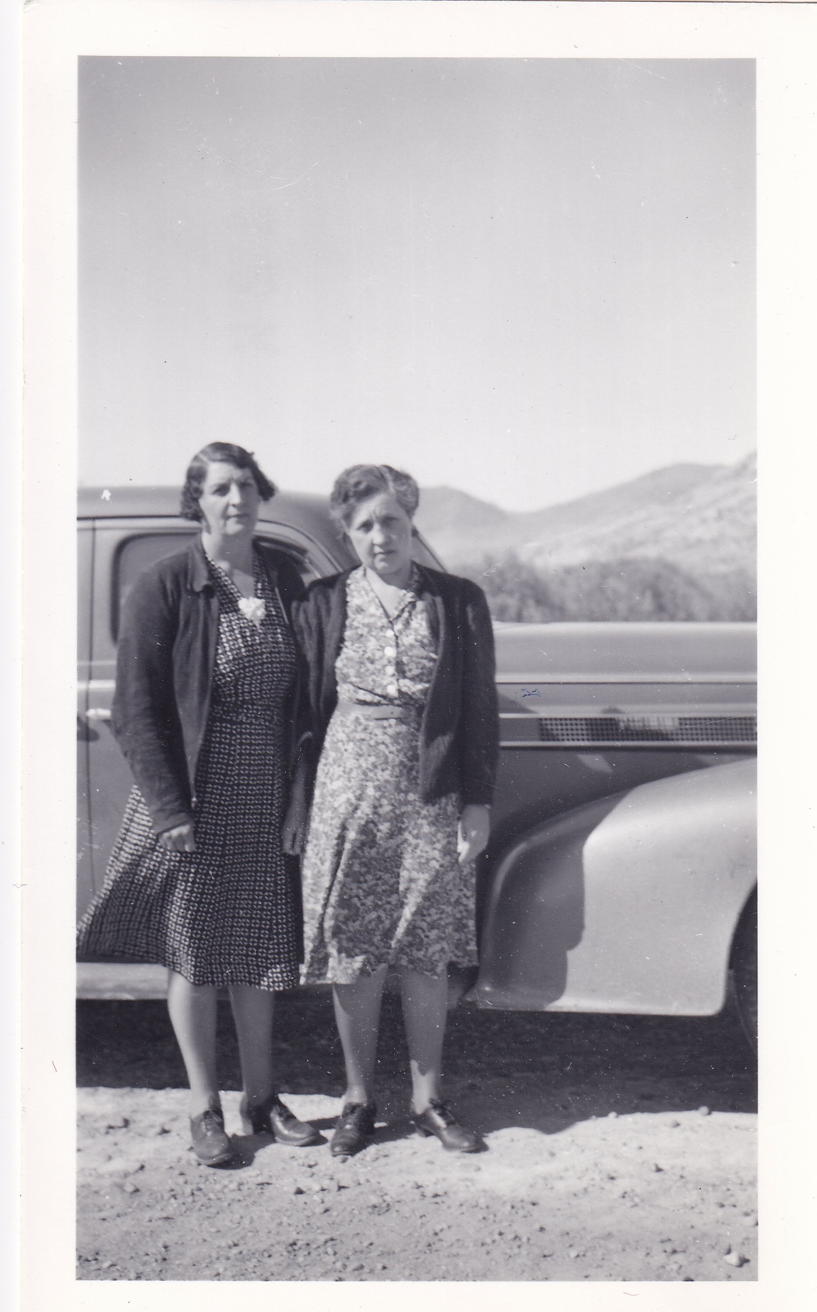 Two women standing outdoors in front of a vehicle with a mountainous landscape in the background. The women are wearing dresses and jackets, and are standing on a grassy and rocky terrain.