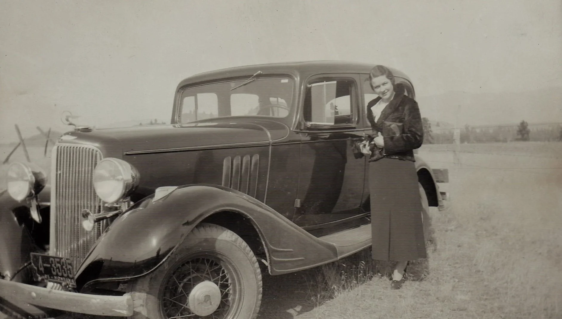 Black and white photo of a young woman standing outside beside a vintage car in an open field, with a fence and trees in the background.