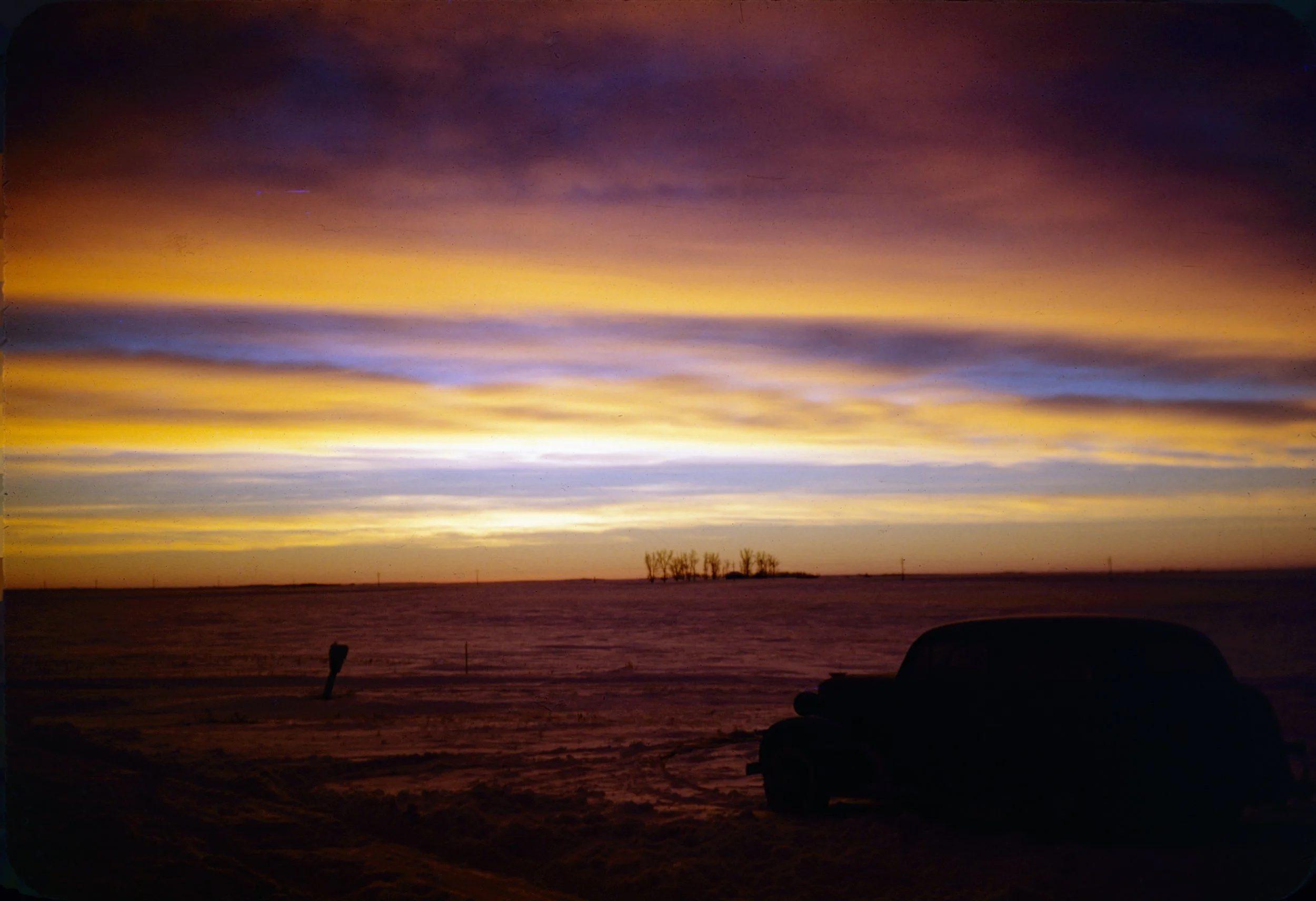 A scenic view of a sunset over a body of water with a few trees and silhouettes of objects in the foreground.