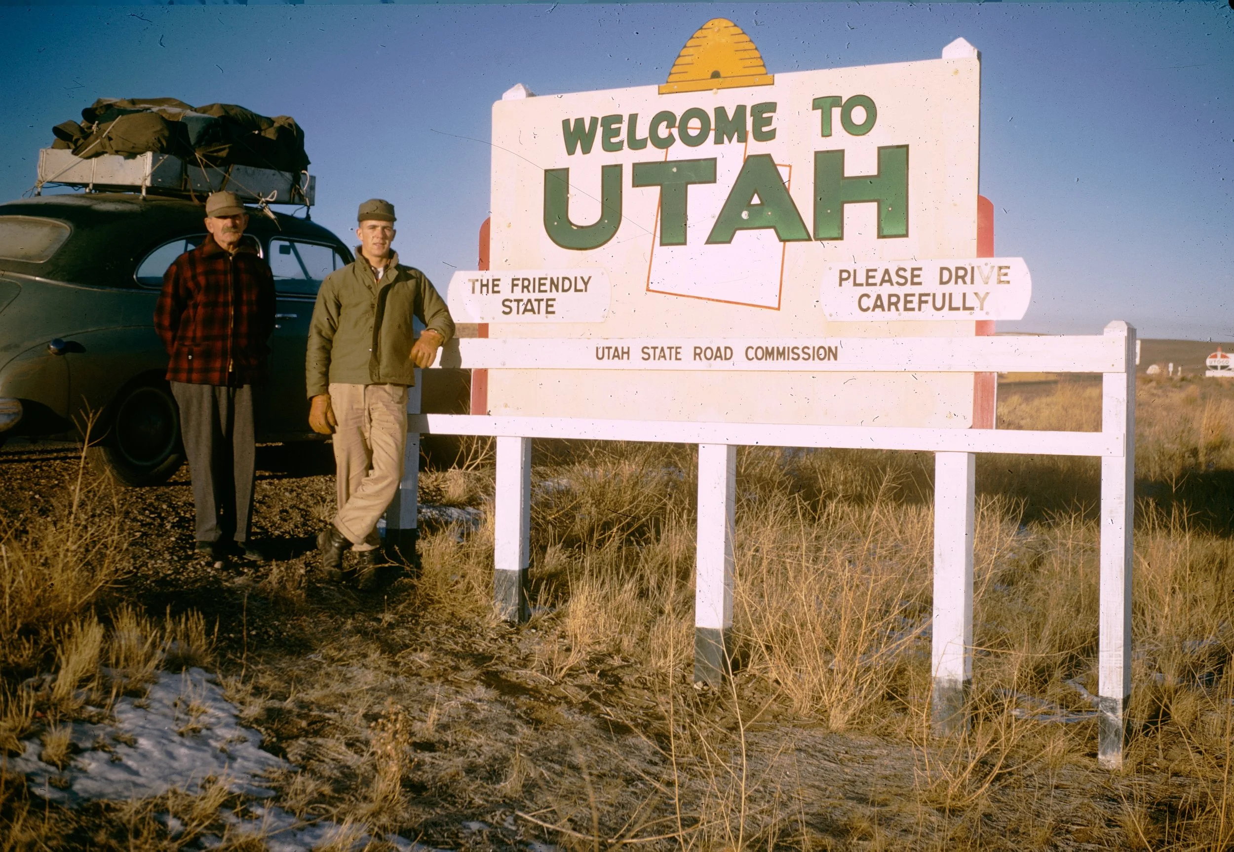 Two men standing in front of a large welcome sign at the Utah border, with a vintage car nearby and an open landscape in the background.