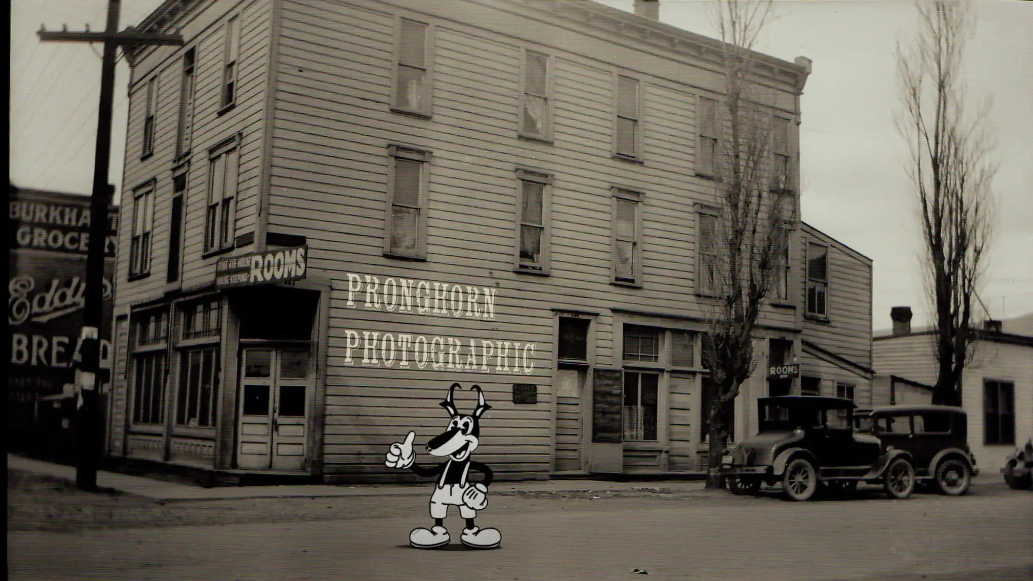 A vintage black and white photograph of a street scene with a three-story building. The building has several windows and a sign that reads 'PRONGHORN PHOTOGRAPHIC.' There are cars parked along the street and a cartoon character of a wolf in shorts and gloves, smiling and pointing upwards, superimposed in the foreground.