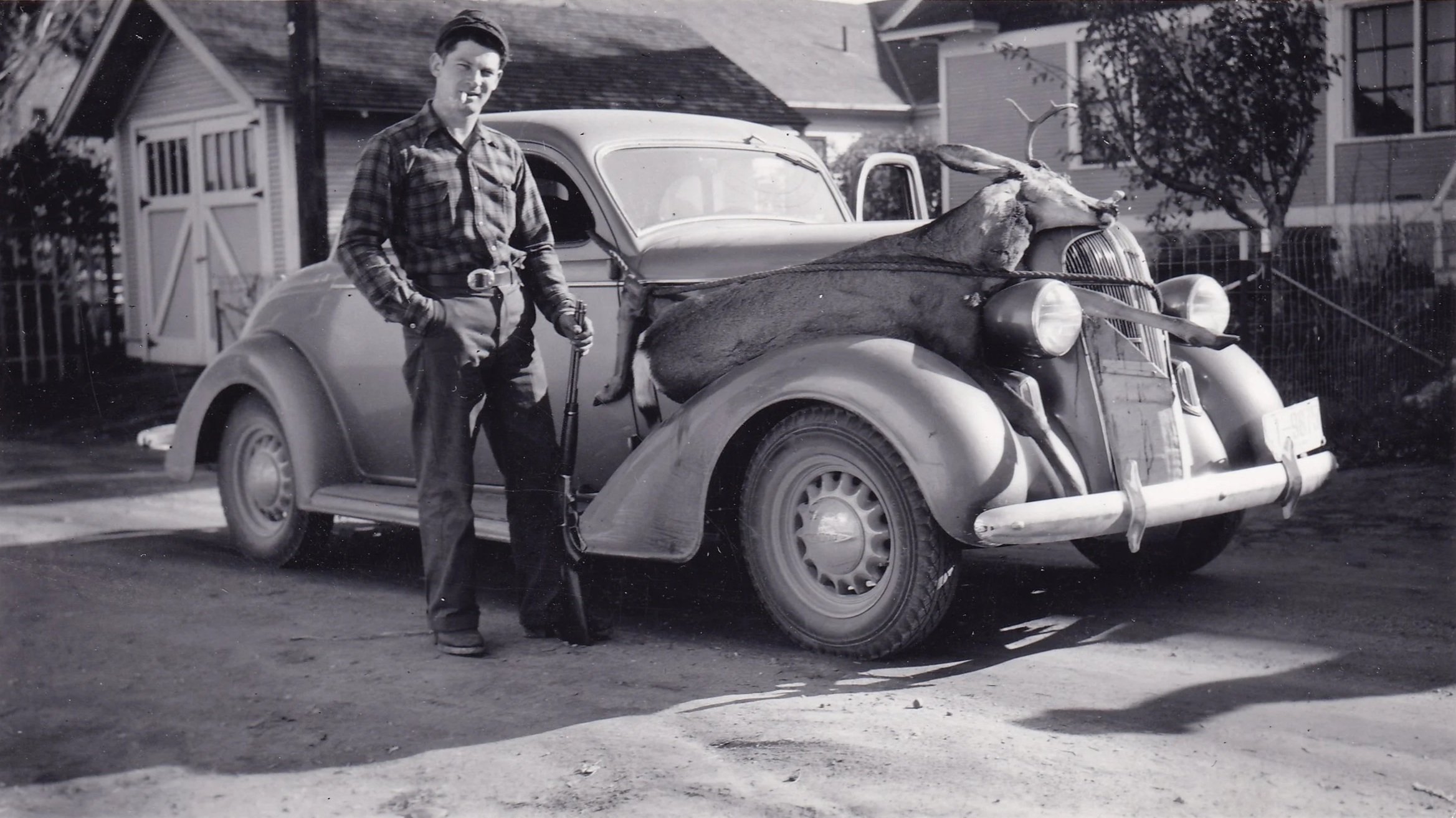 A young man in plaid shirt and slacks standing beside a vintage car with a dead deer on the hood, in a residential neighborhood.