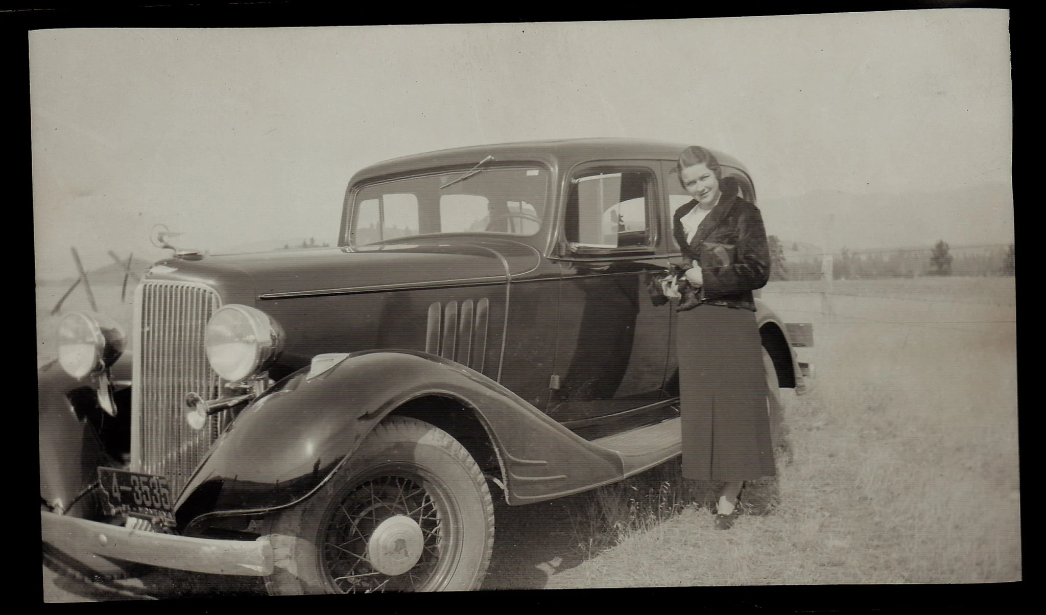 Black and white photograph of a woman smiling and standing next to a vintage car in an open field.