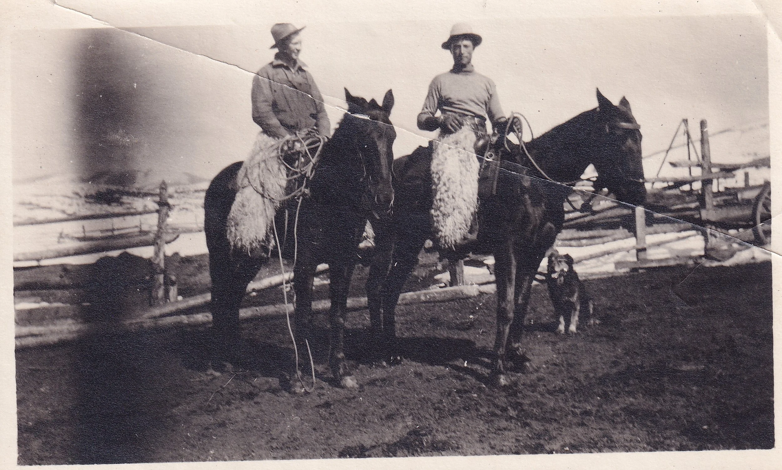 Black and white photograph of two people on horseback, one holding a dog on a leash, standing on a dirt path with snow-covered landscape and wooden fences in the background.