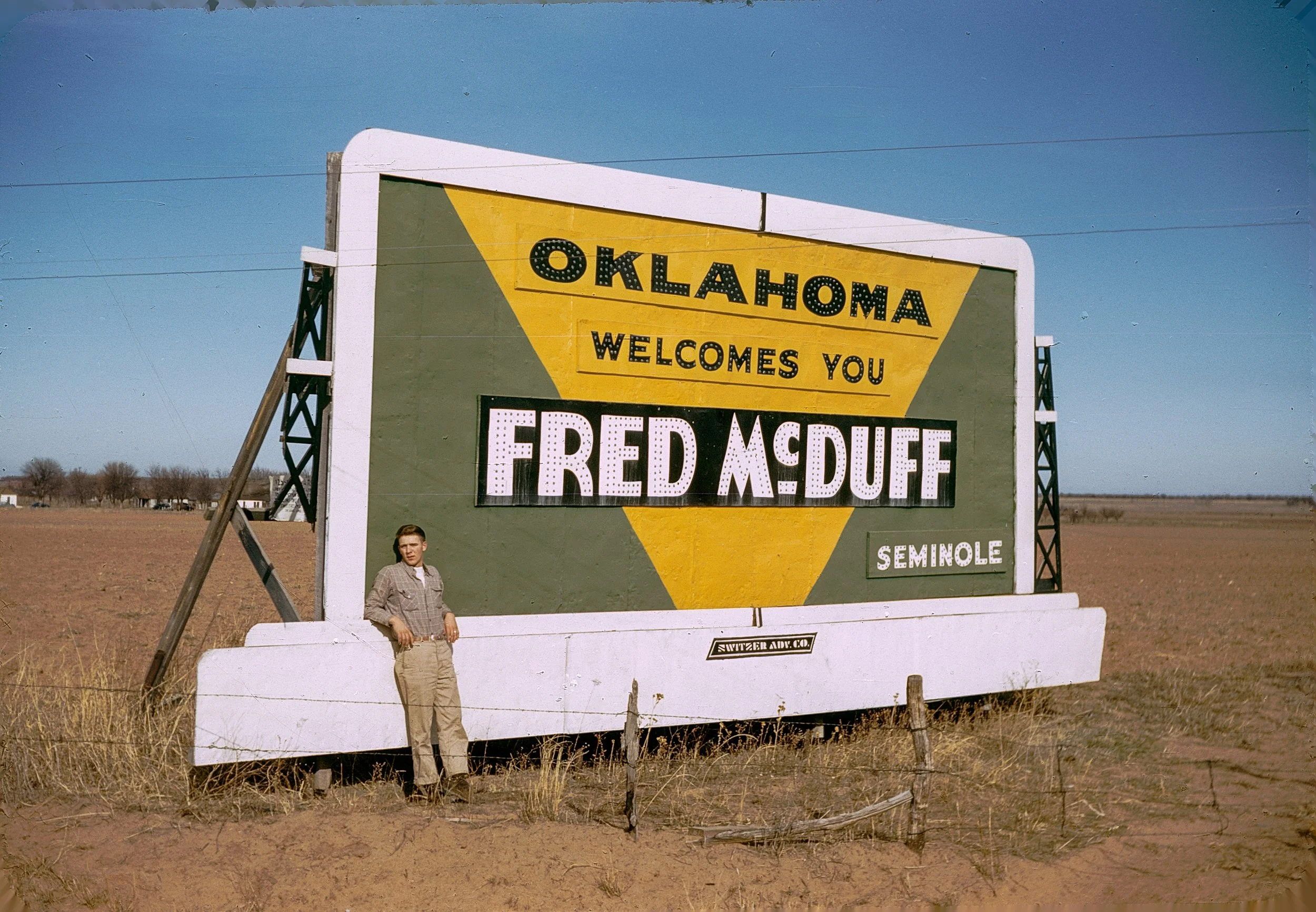 A large sign on the side of a rural highway reads "Oklahoma Welcomes You" and promotes Fried Muffin and Seminole. A man is standing next to the sign in a plaid shirt and khaki pants, with a wide open field in the background.