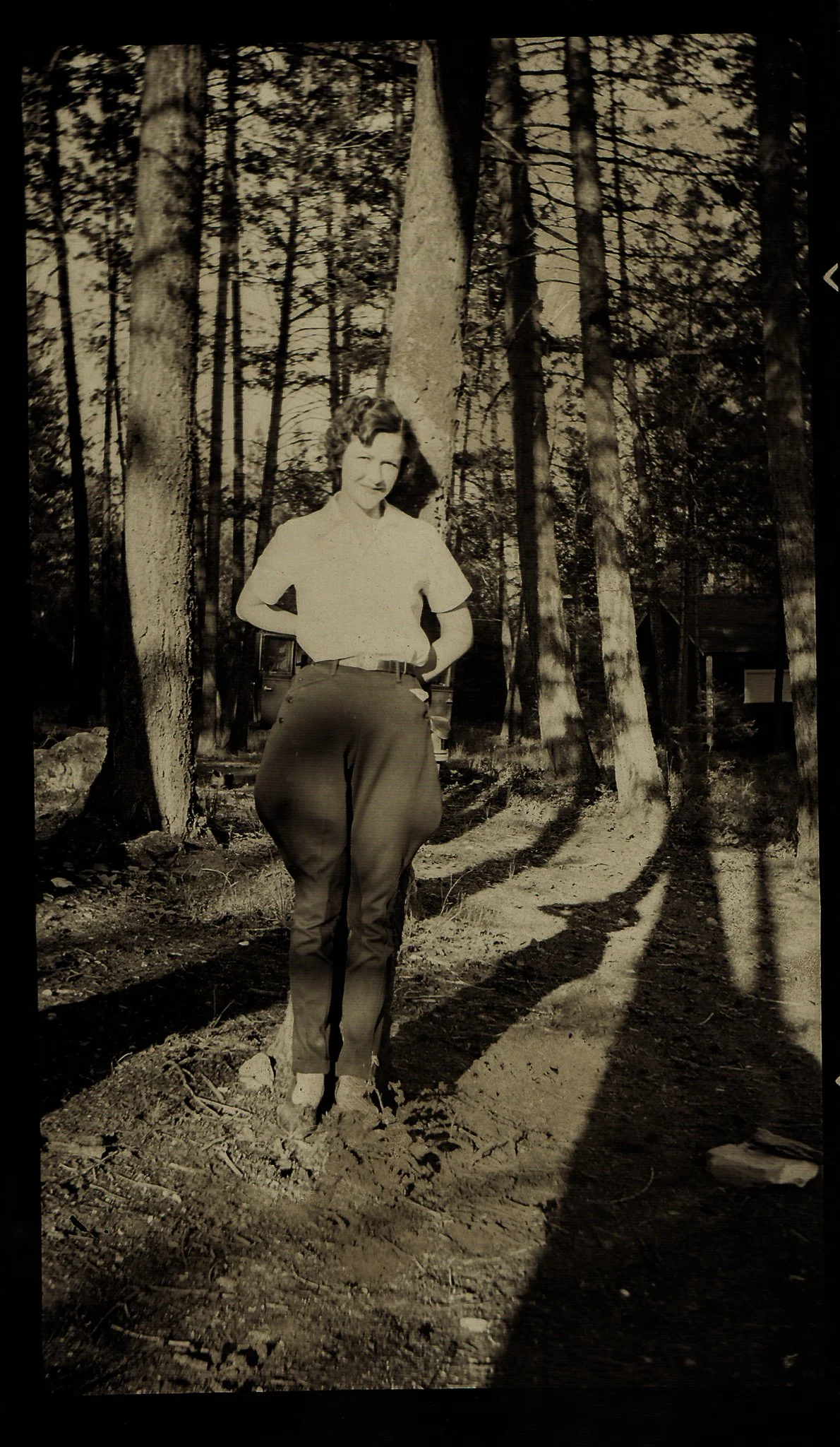 A woman standing on a dirt path in a forest, with tall trees casting long shadows. She has her hands on her hips and is smiling.