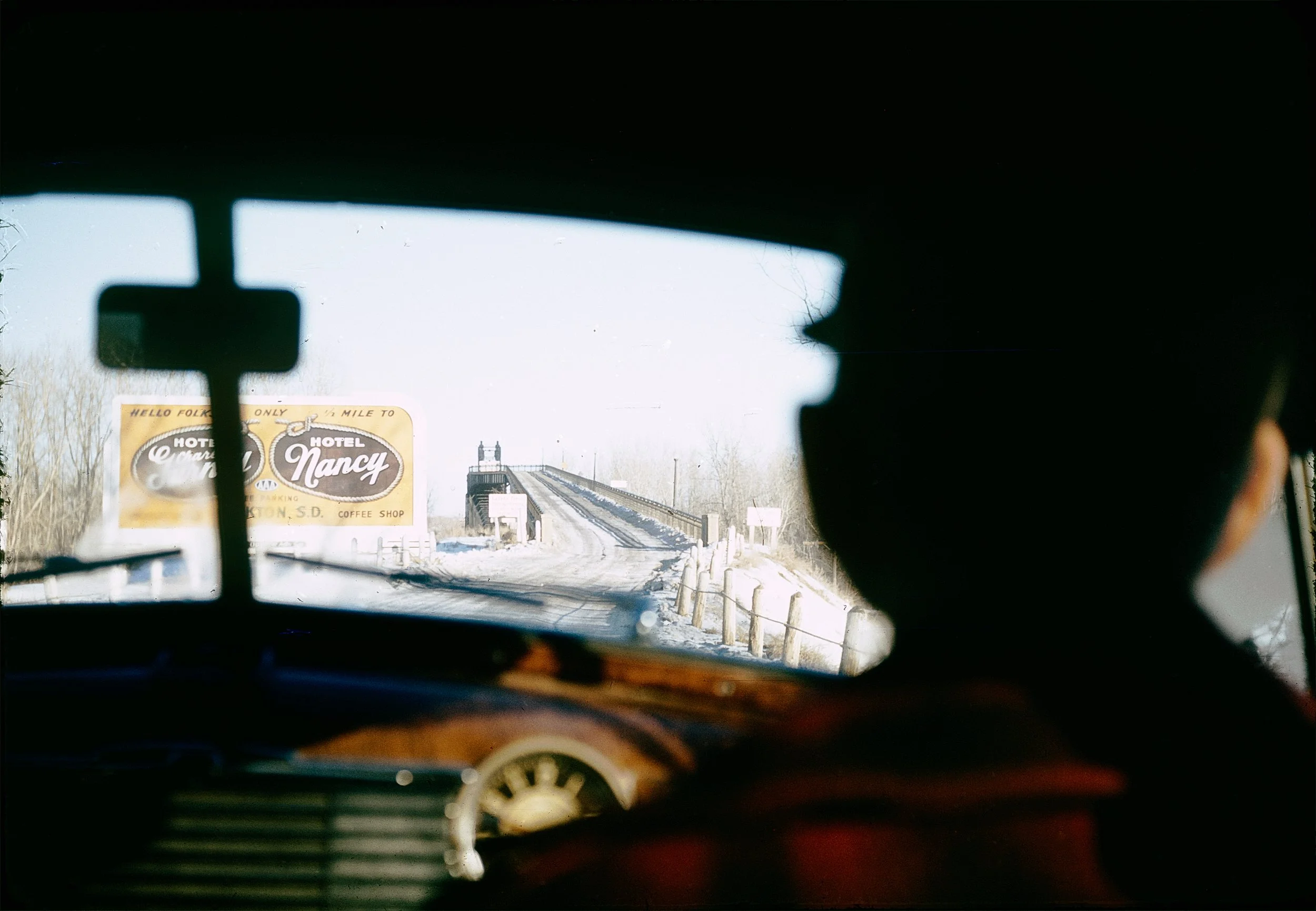 View from inside a vehicle looking out through the windshield, showing a bridge, a billboard for a hotel, and a snowy landscape outside