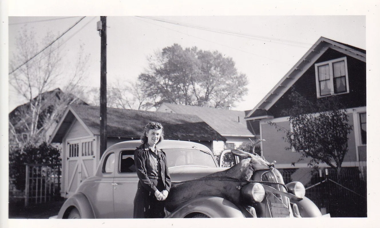 A woman standing next to a vintage car with a taxidermy animal on the hood in a suburban neighborhood yard.
