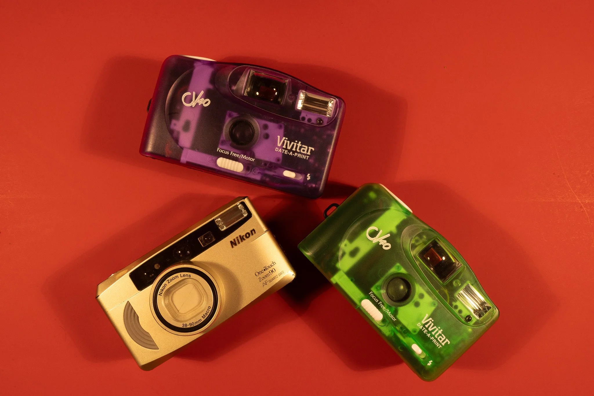 Three vintage cameras placed on a red surface, including a silver Nikon compact camera and two colorful Vivitar instant cameras, one purple and one green.