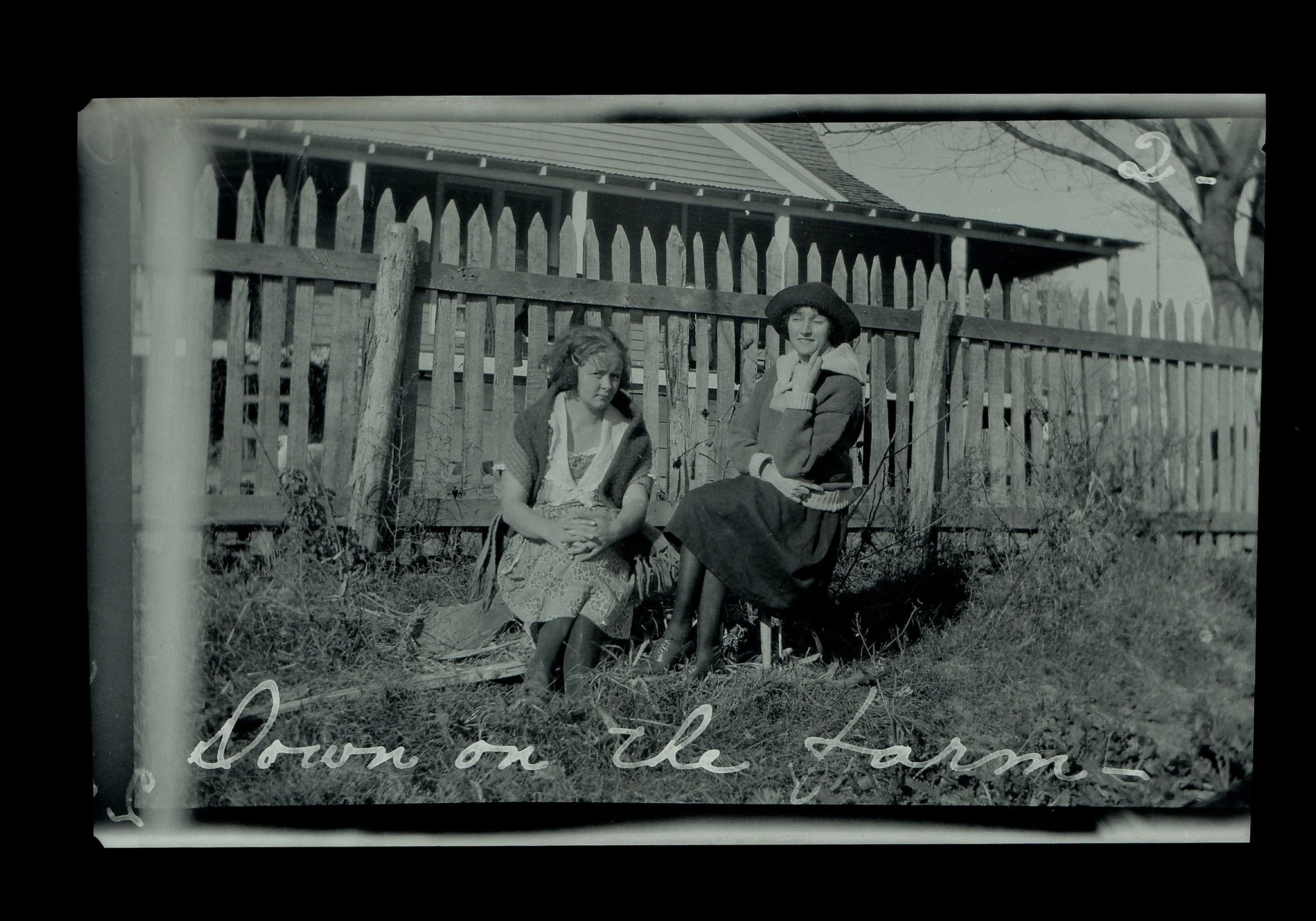 A black and white photograph of two women sitting on a wooden fence outside a house with a sloped roof. One woman is wearing a hat and light-colored sweater, while the other has curly hair, a darker jacket, and a patterned skirt. The background inclu