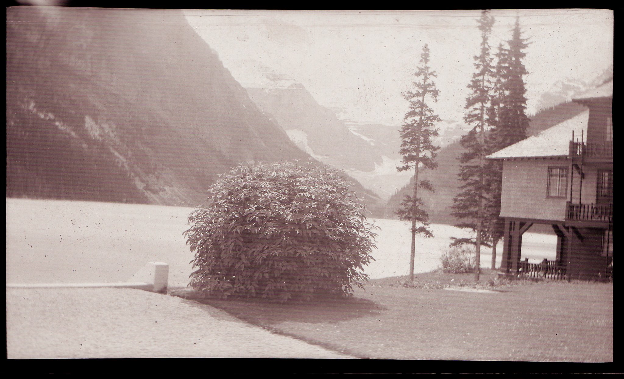 A black and white photo of a house with a porch and balcony, surrounded by tall trees, mountains, and snow in the background.