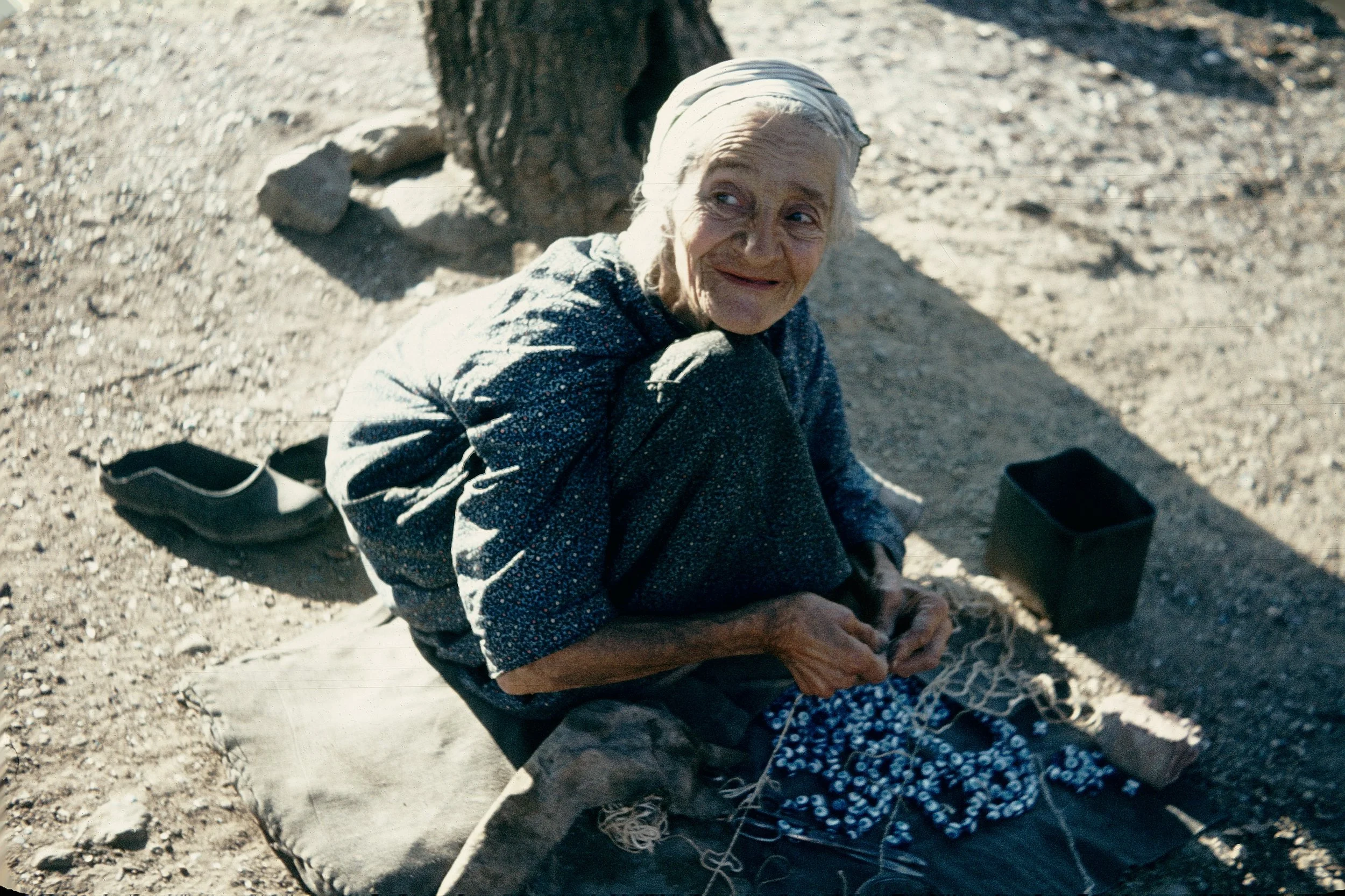 An elderly woman with gray hair, sitting on the ground outdoors in a dirt area, smiling and looking to the side. She is wearing a headscarf and patterned clothing, with some personal items placed nearby.