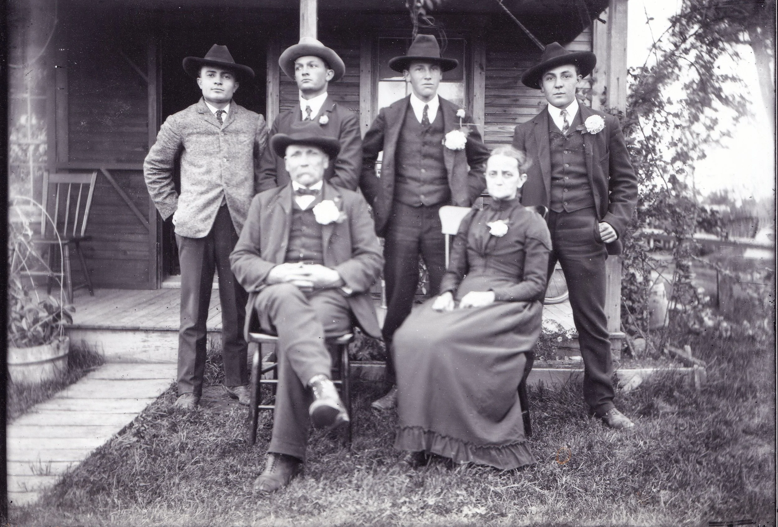A black and white photo of five people, four standing in the background and one seated in the foreground, outside a rustic wooden building with plants and a small porch. All individuals are dressed in old-fashioned clothing, likely from the late 19th