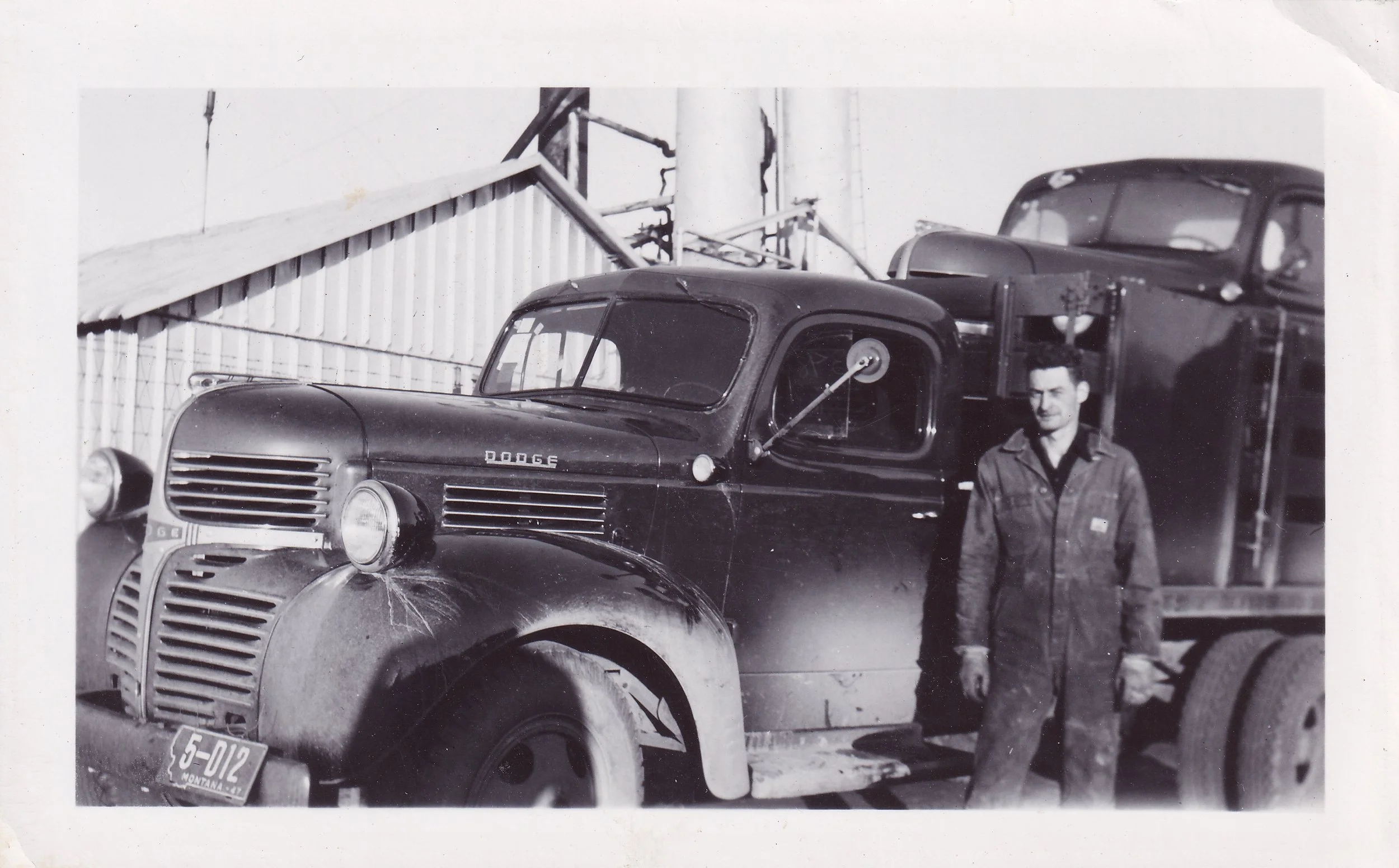 Black and white photo of a man standing next to a vintage Dodge truck with a split windshield, parked outside a building with a metal roof. There are two cars on the truck's trailer, one partially visible.