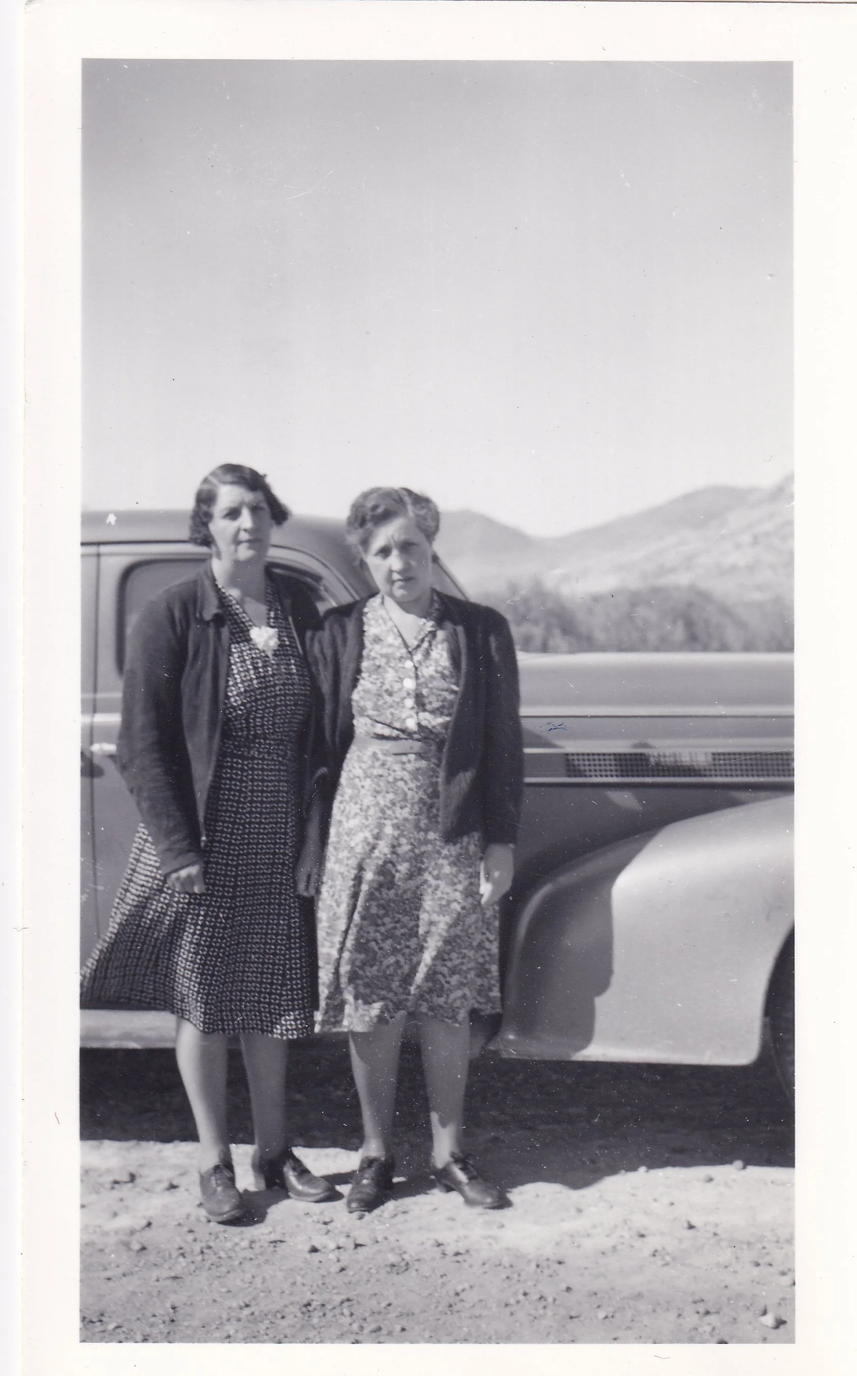 Two women standing outdoors beside a vintage truck, with mountains in the background, dressed in mid-20th century clothing.