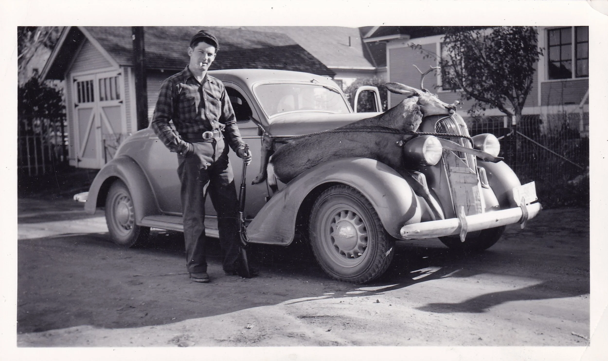 Black and white photo of a young man in checkered shirt and jeans posing next to a vintage car with a dead deer strapped to the front grille.
