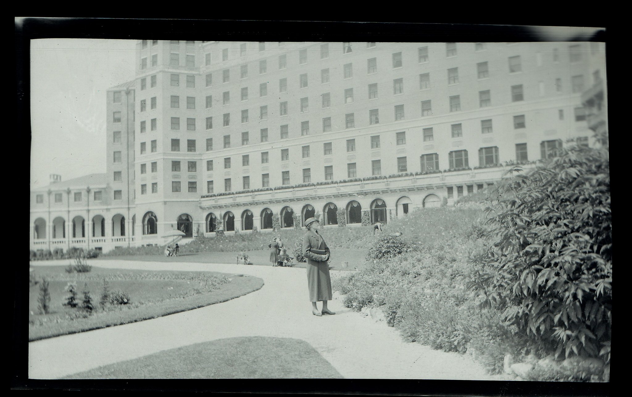 Black and white photo of a large hotel building with multiple windows and balconies, seen from a landscaped park with a winding path, shrubs, and trees. In the foreground, a woman wearing a coat and hat stands near some bushes, with a few children an