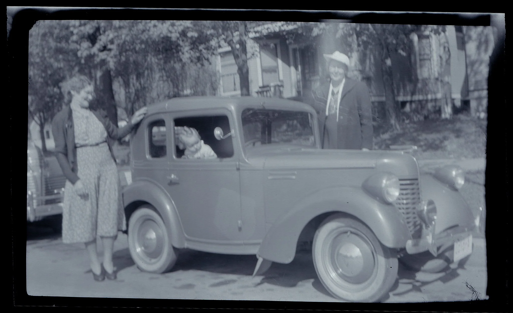 Black and white photo of a woman and a child near a vintage automobile on a city street, with buildings and trees in the background.