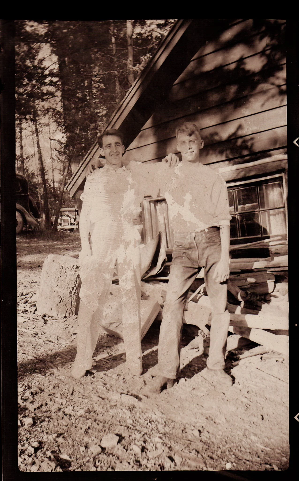 Black and white photo of two young men standing outside in front of a wooden building, posing with their arms around each other. They are on a rocky ground, and there are trees and a vintage car in the background.