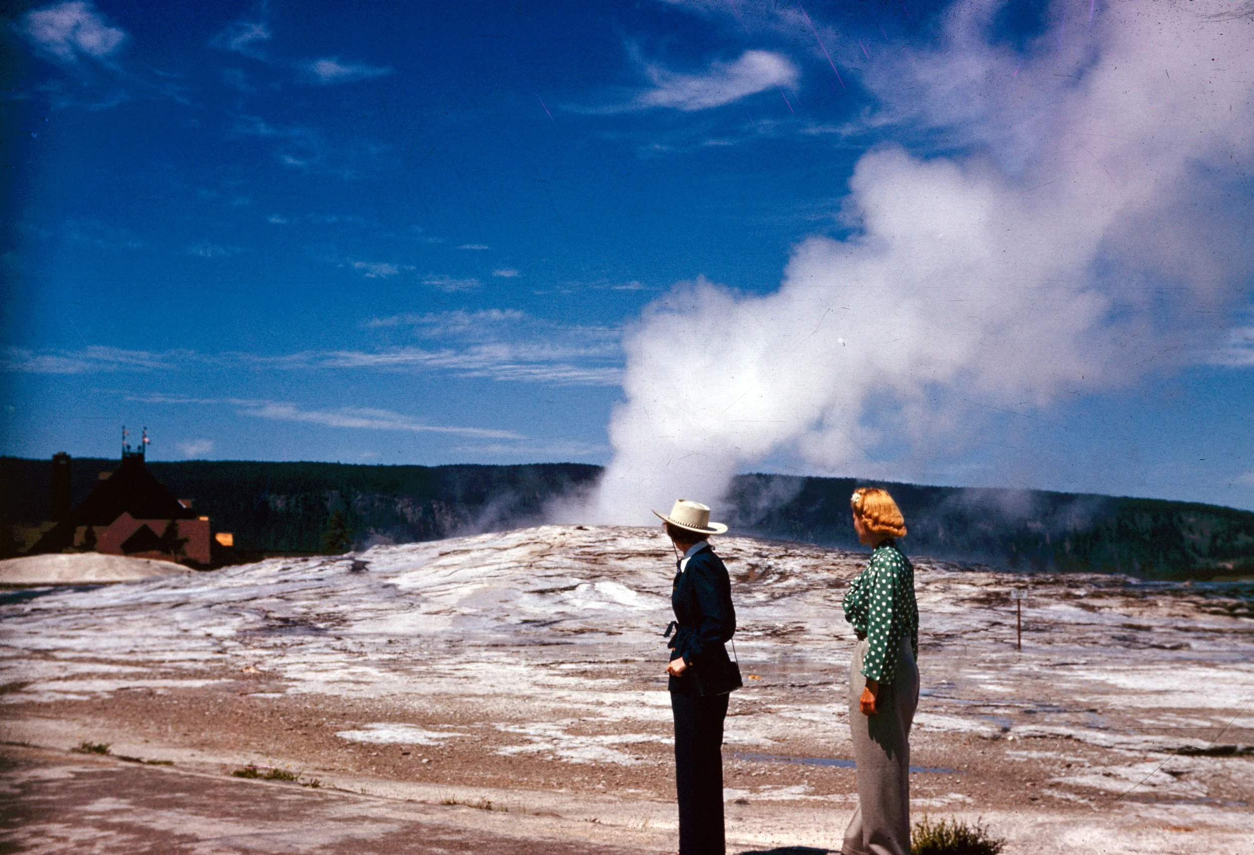 Two people, a man and a woman, walking on a rocky landscape with steaming geysers and volcanic activity in the background, under a blue sky with clouds.