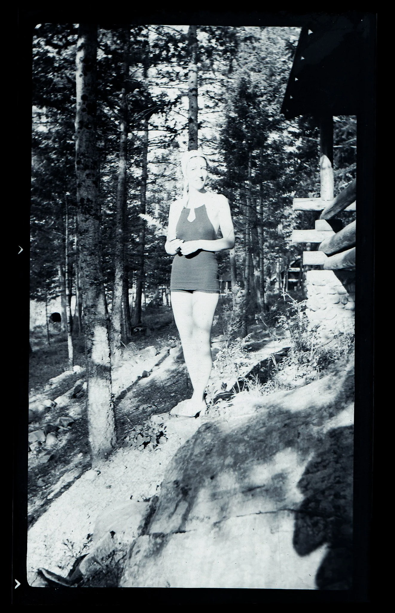 A black-and-white photo of a woman standing outdoors in a forested area with trees and rocks, wearing a sleeveless top and shorts, with a cabin or structure partially visible to the right.