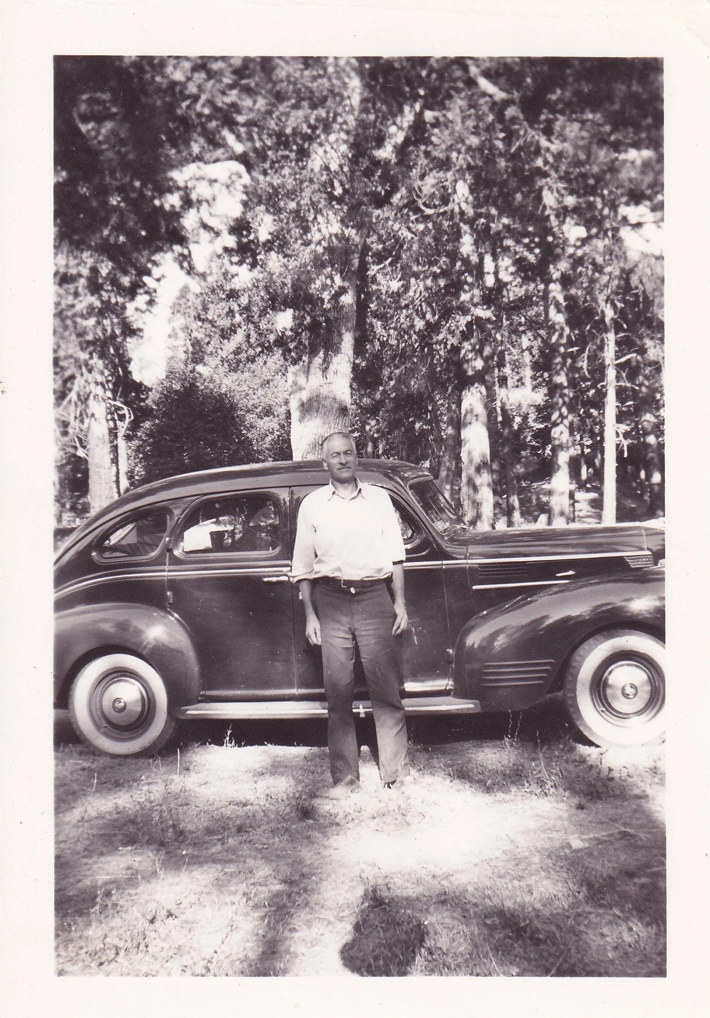 Black and white photograph of a man standing next to a vintage car outdoors with tall trees in the background.
