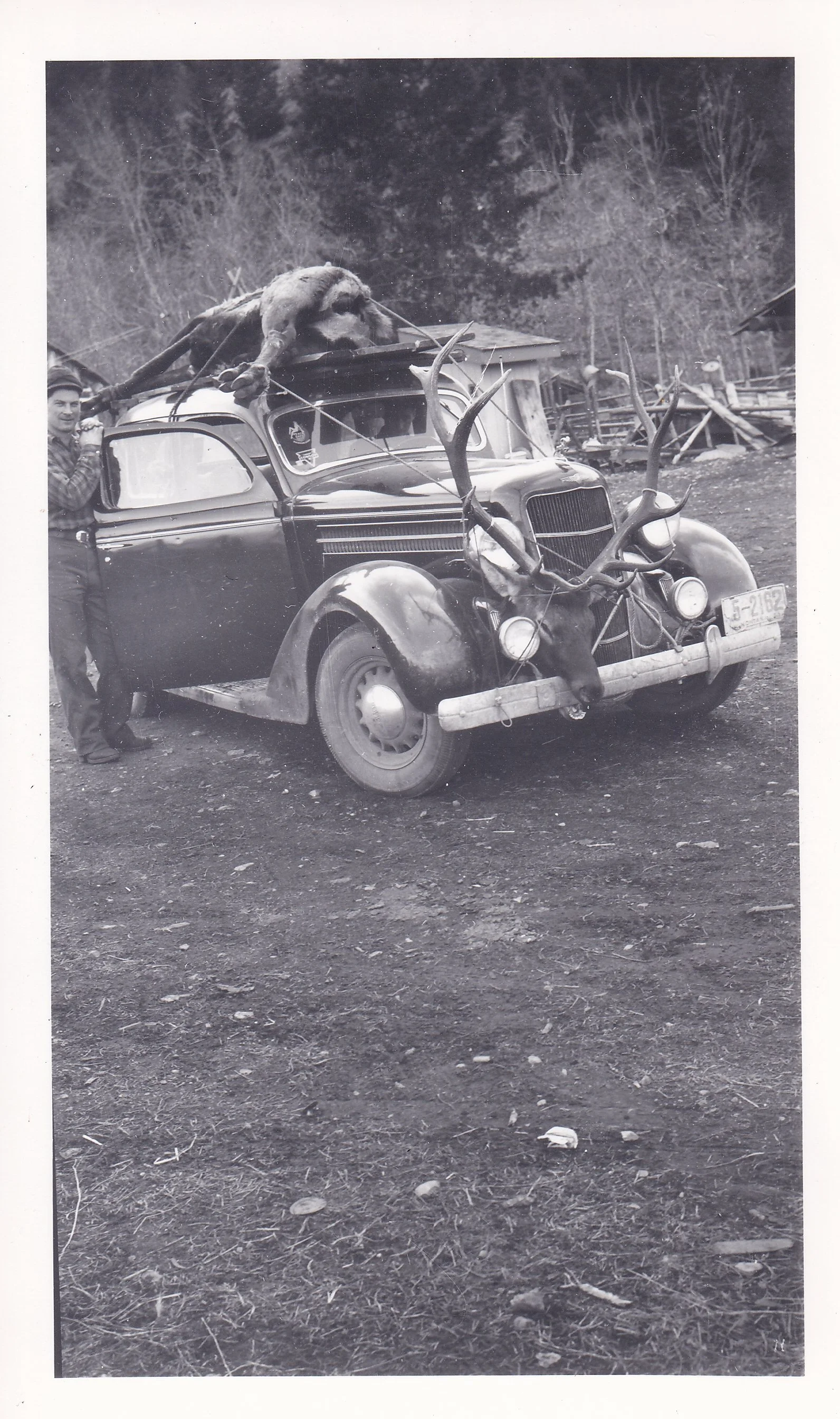 Black and white photo of a man standing next to a vintage car decorated with deer antlers on the front and a large animal on top, outdoors with trees and wooden structures in the background.