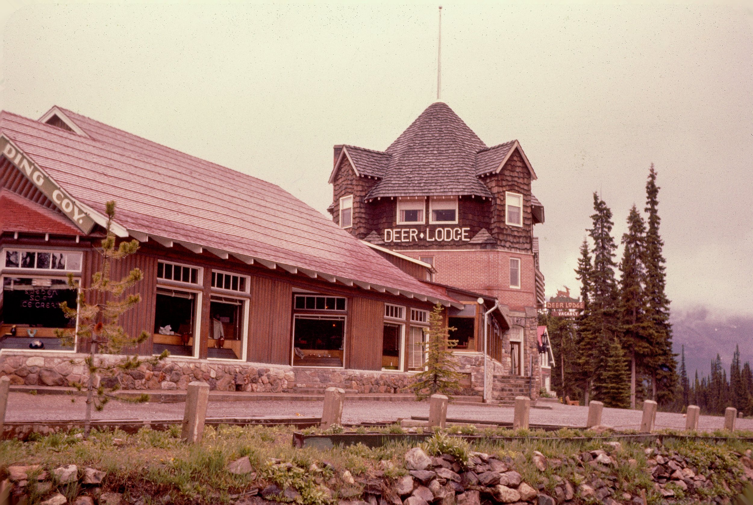 A rustic building labeled 'Deer Lodge,' with a tower-like structure and a weather vane on top, surrounded by tall trees and set against a mountainous background.