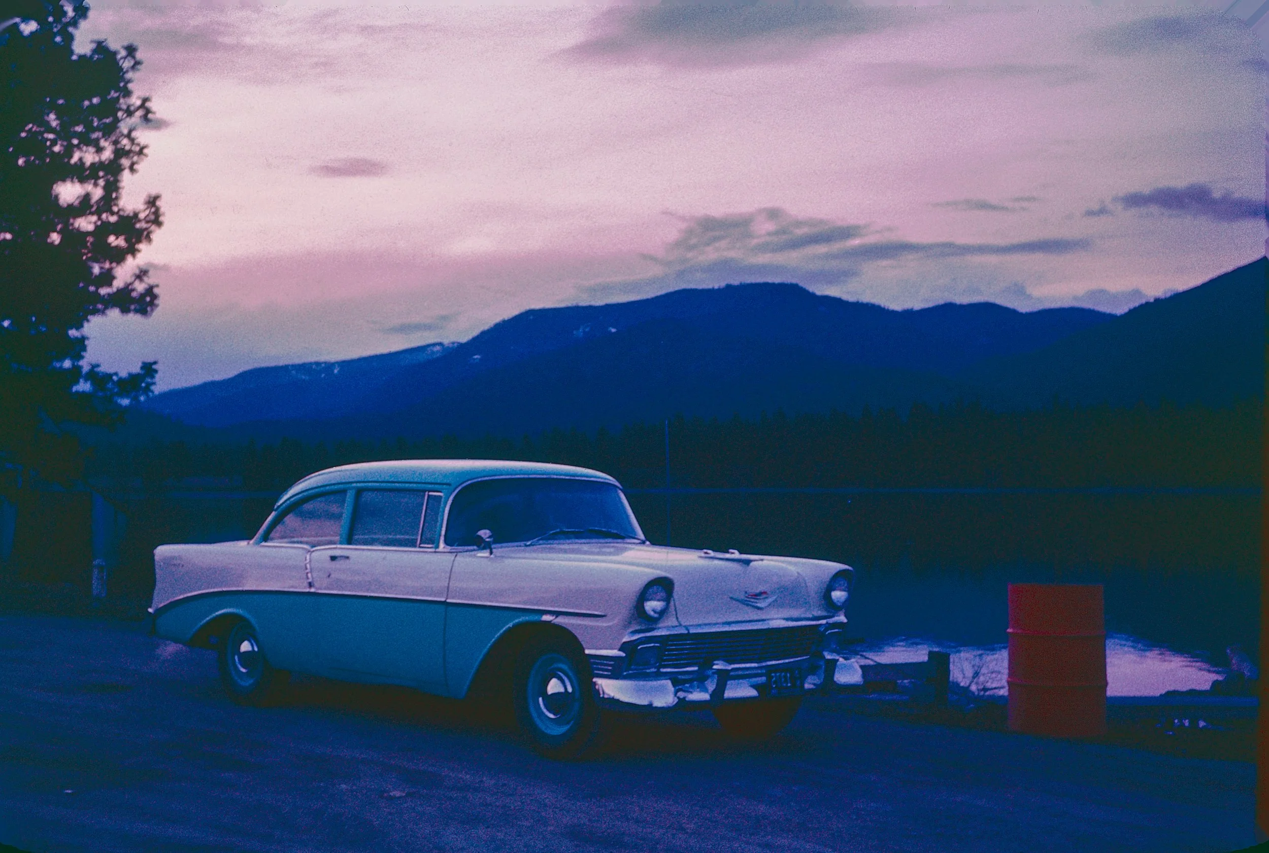A vintage white car with a rounded body shape parked at a scenic location with mountains in the background during dusk or dawn.