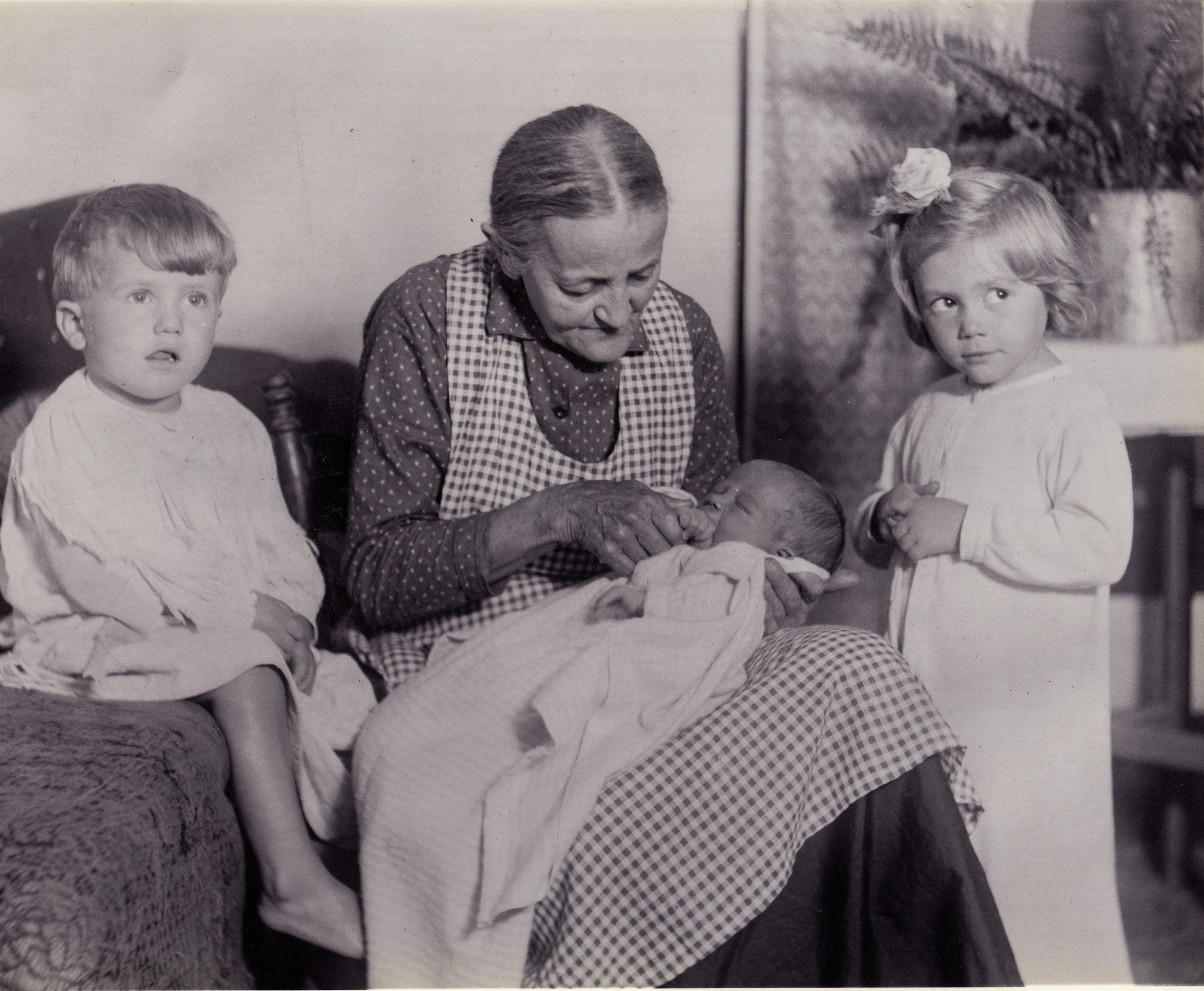 Black and white photo of two young children and an elderly woman sitting together in a room, with the woman sewing or knitting.