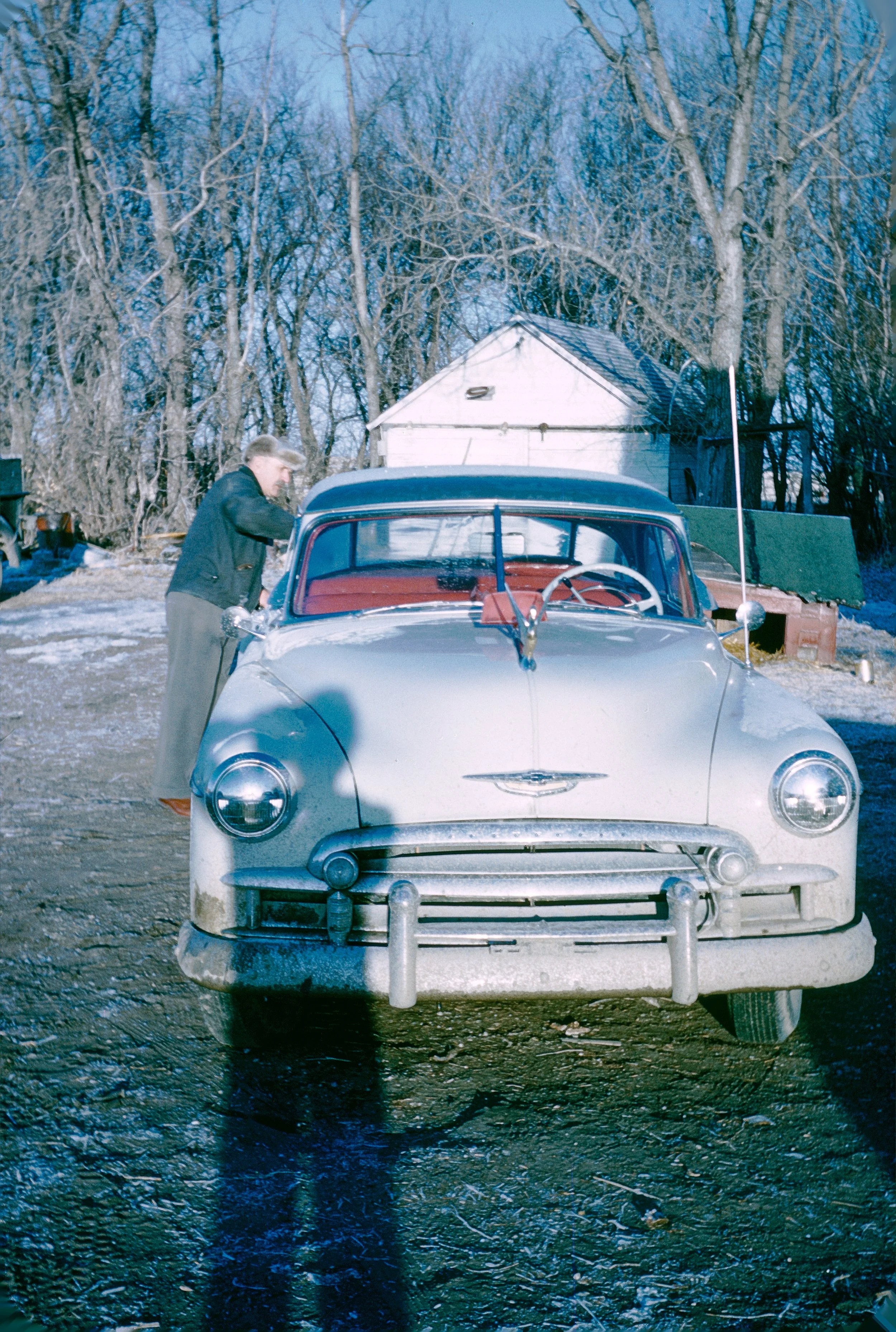 A man in a dark jacket working on a vintage gray car with a red interior, parked in a rural backyard with leafless trees and a small shed, during winter.