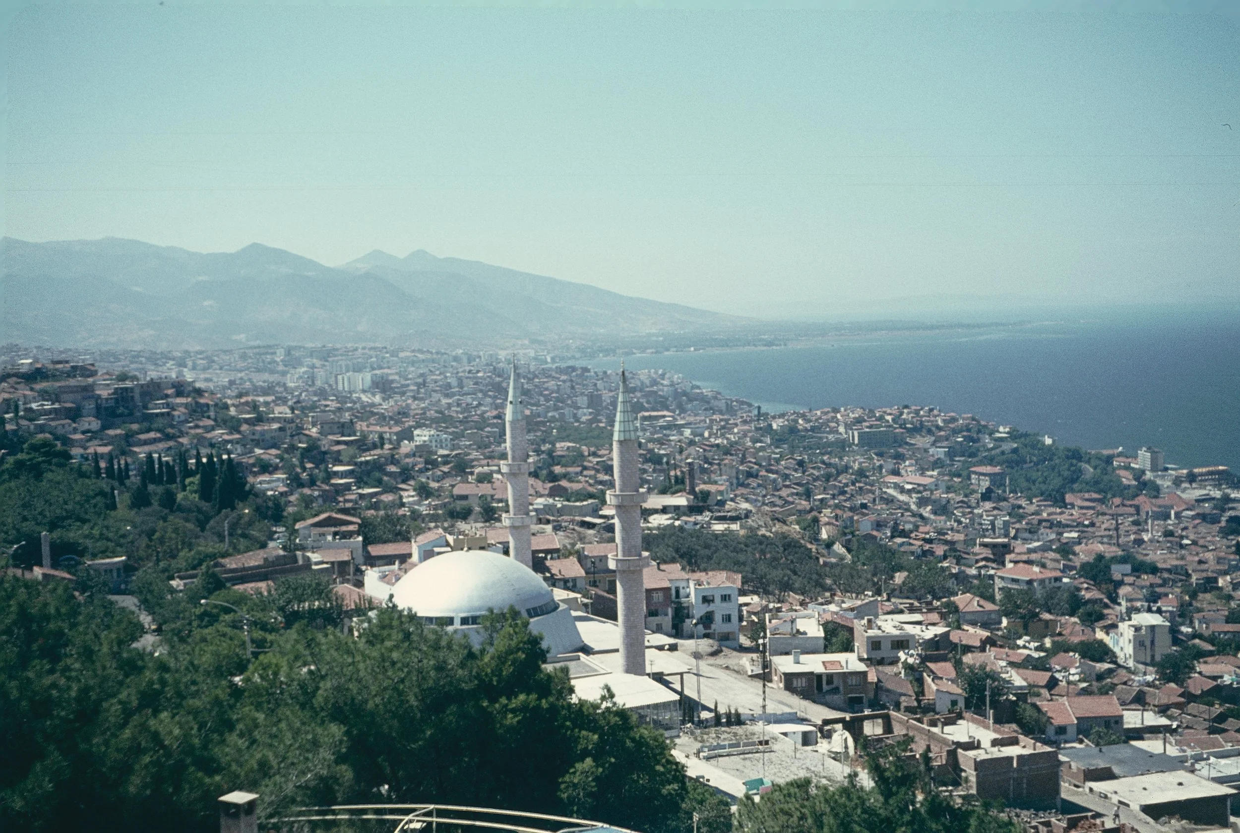 A cityscape with many houses, two prominent church steeples, green trees in the foreground, mountain range in the background, and the coast in the distance under a clear blue sky.