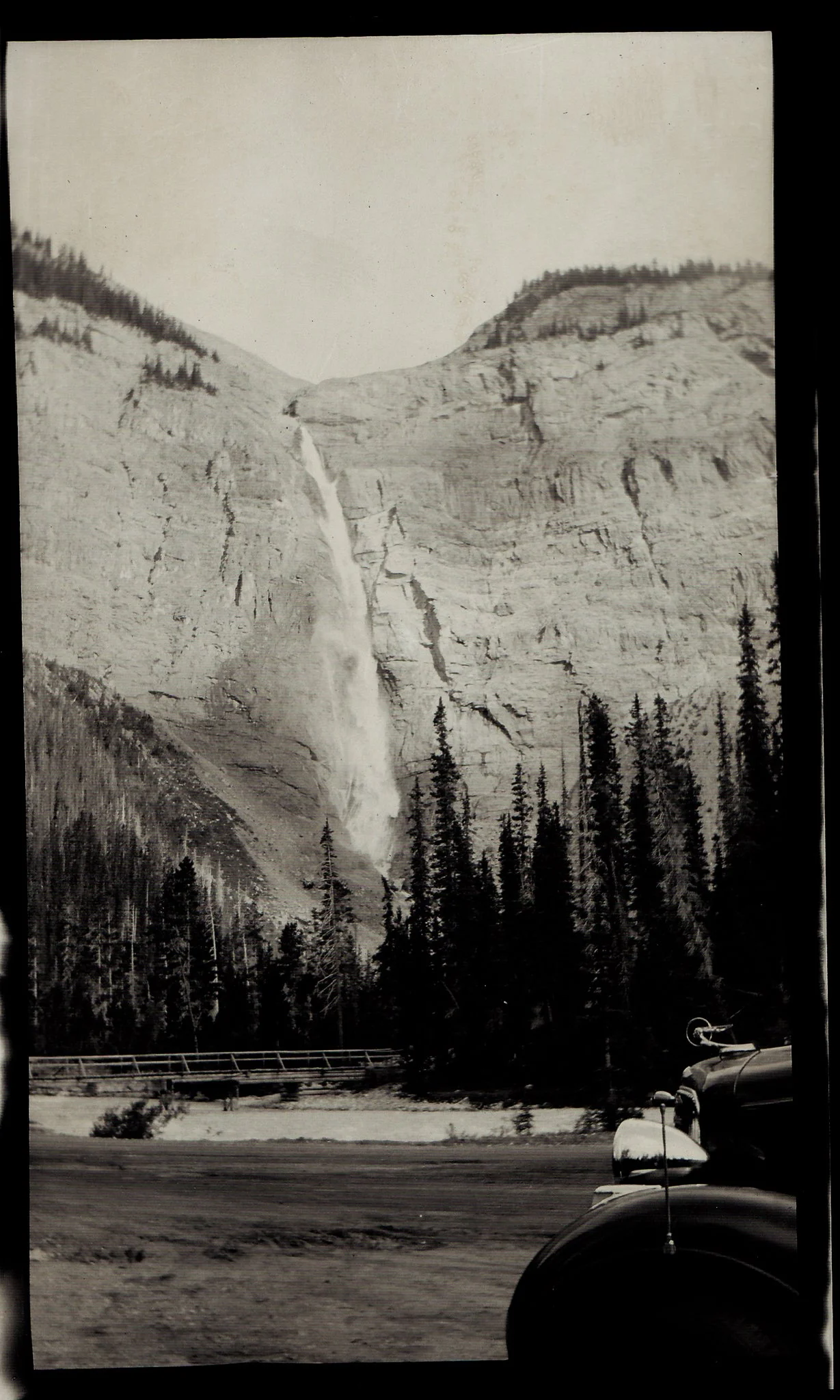 Black and white photograph of a mountainous landscape with a waterfall, evergreen trees, and a road with parked cars in the foreground.