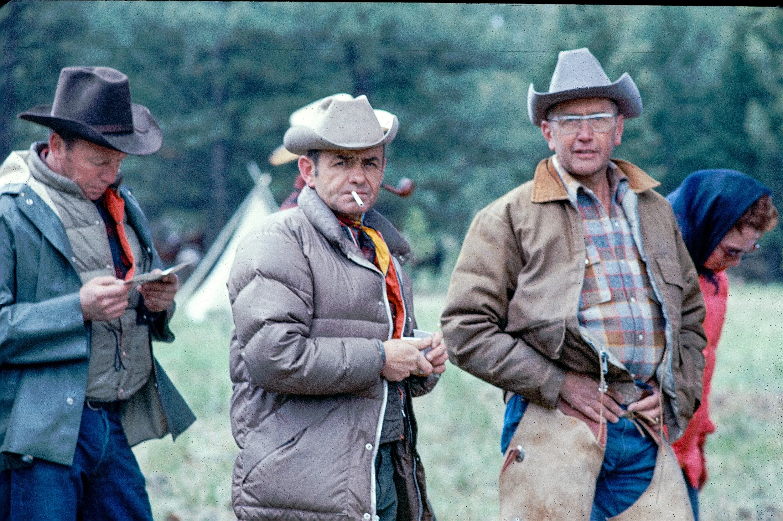Four men wearing cowboy hats outdoors, two of them holding papers, one smoking a cigarette, and a woman partially visible in the background.