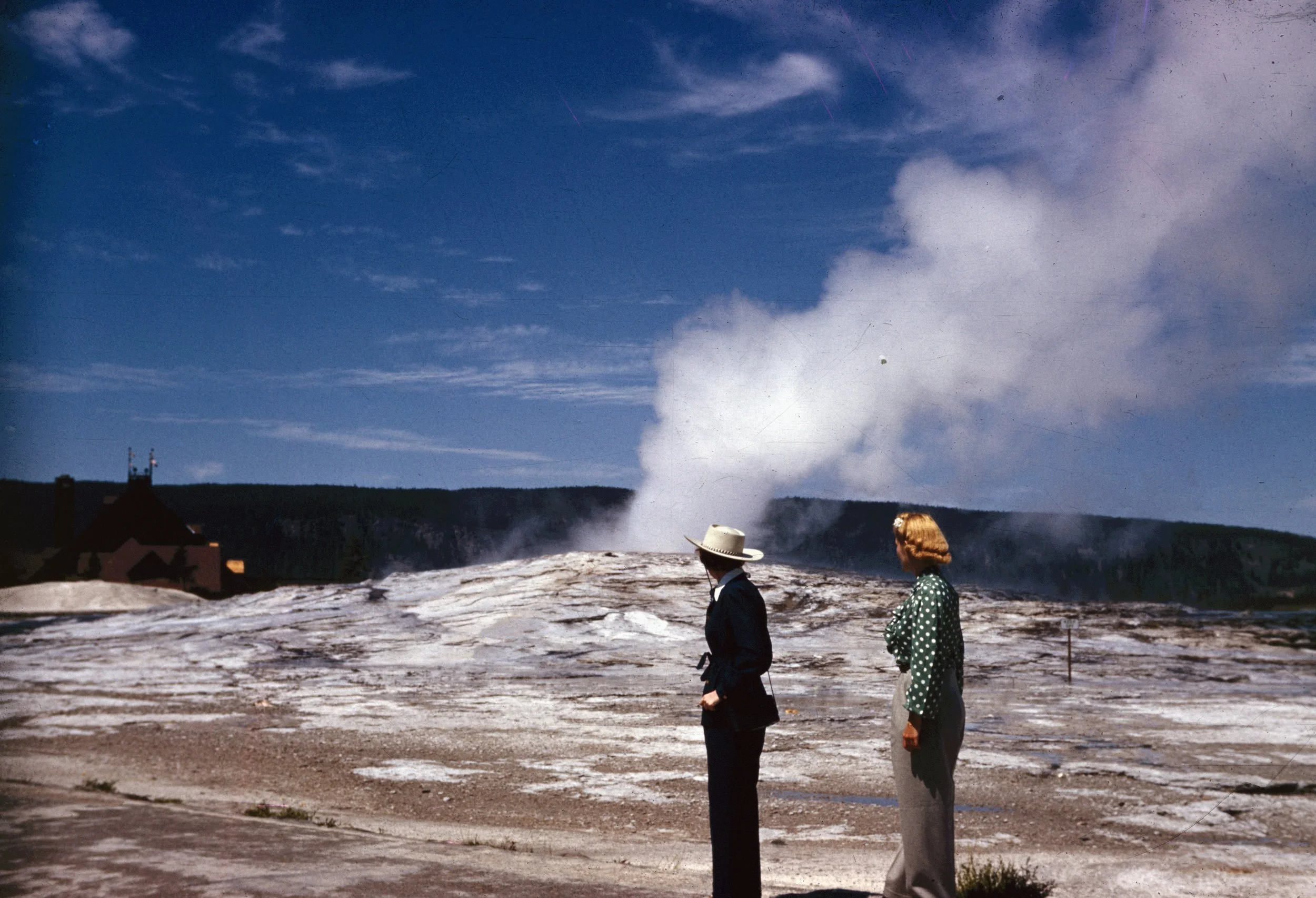 Two children, a boy and a girl, walk on a rocky terrain with a smoky geyser erupting in the background under a partly cloudy sky.