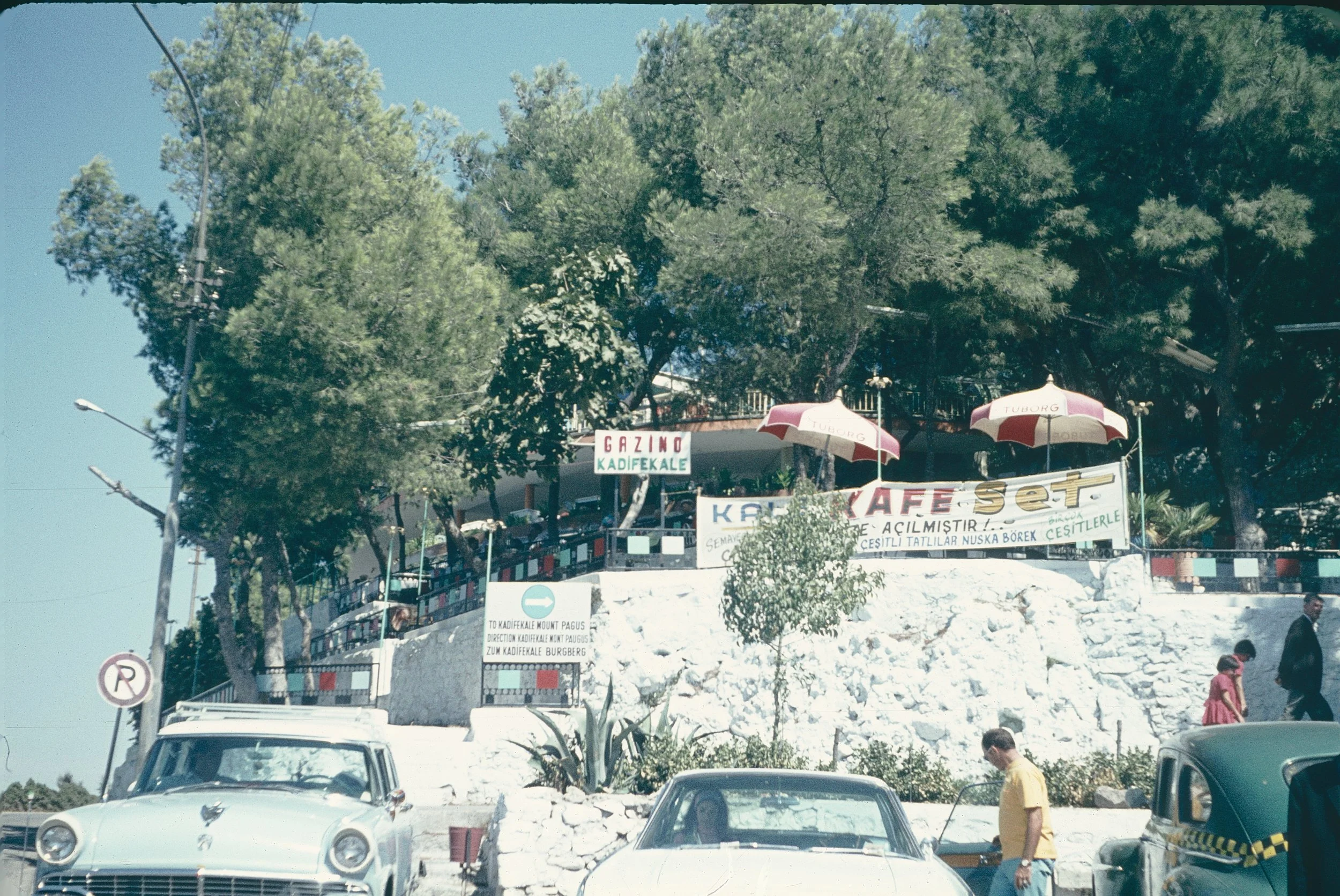 Street scene with vintage cars parked in front of a hill with green trees, signs, and umbrellas with the word 'Tuborg' on them.