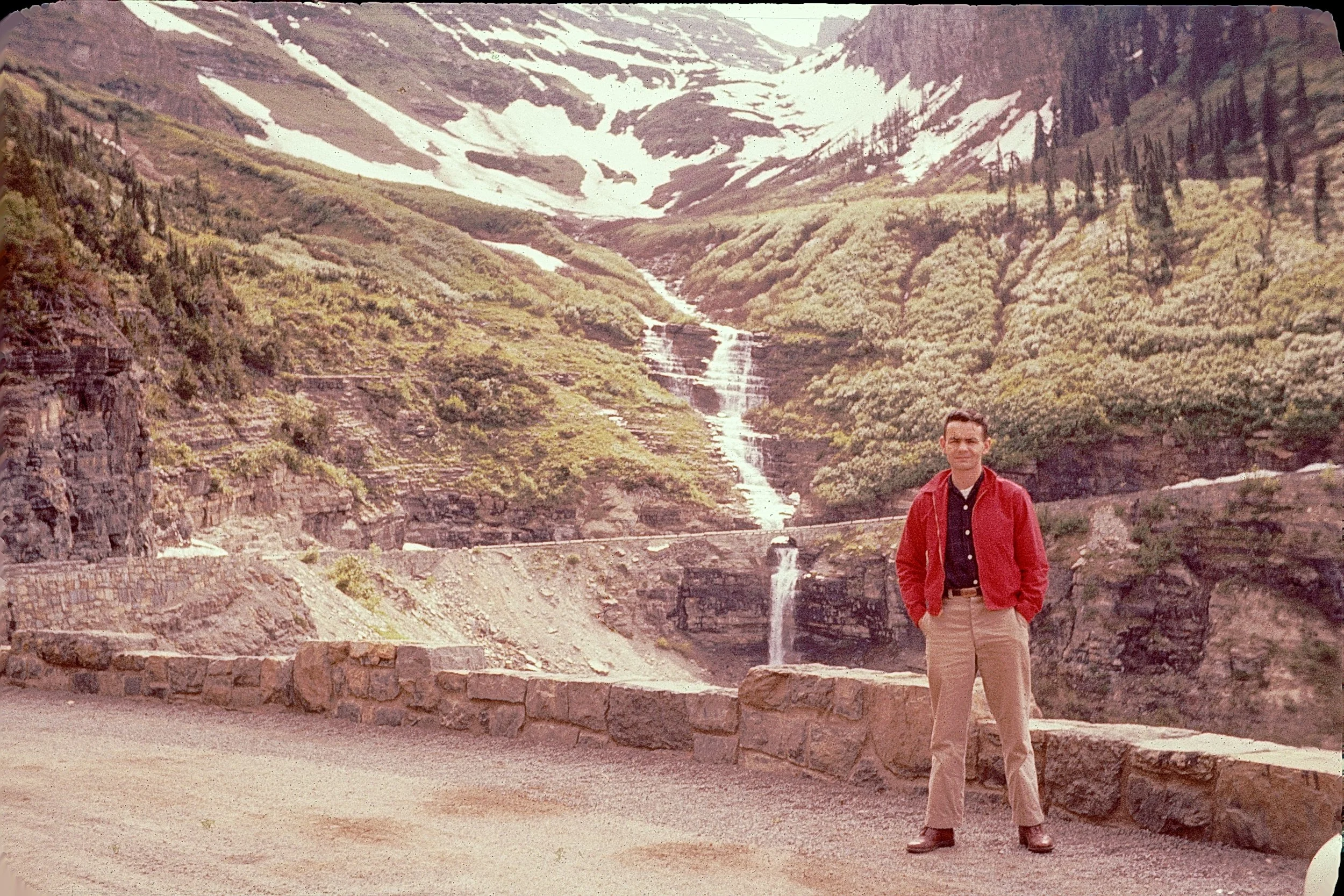 Man in a red jacket and beige pants standing on a stone platform by a mountain river with a waterfall, surrounded by green vegetation and snowy mountain peaks in the background.