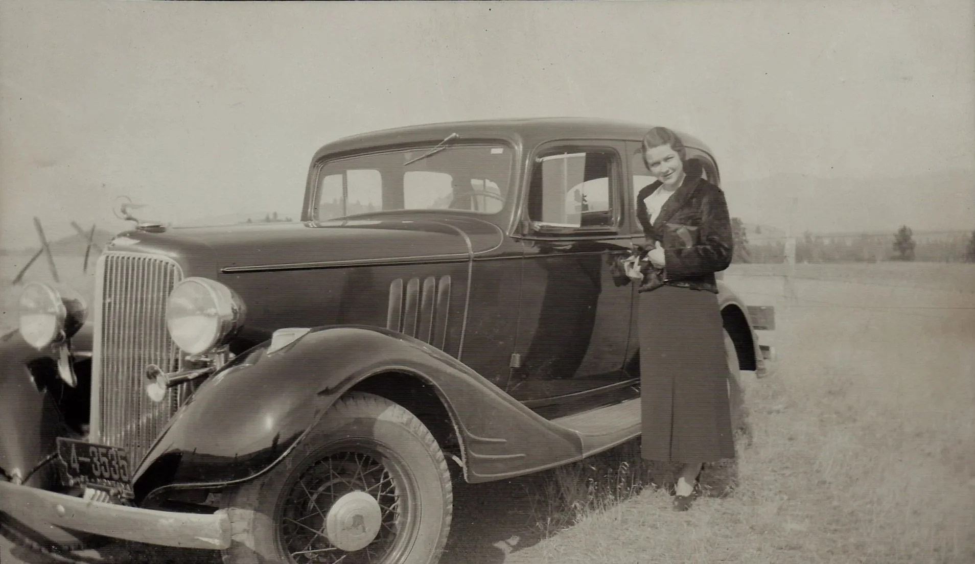A vintage black-and-white photo of a woman standing next to a classic automobile, holding a bouquet of flowers. The car is parked on a grassy field with a fence and some trees in the background.