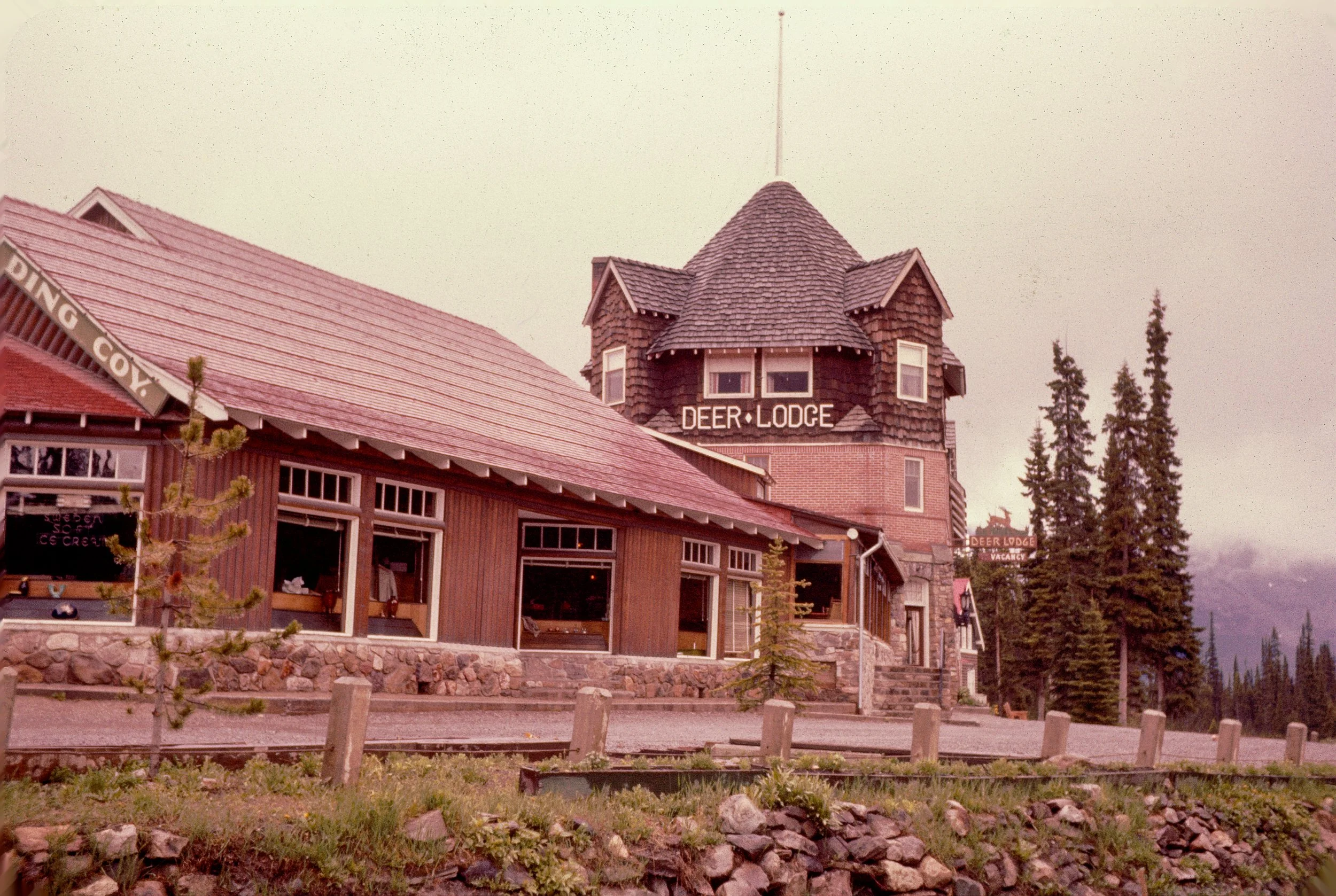 A wooden building with a tower labeled 'Deer Lodge' and a sign that says 'Vancouver.' The building has a shingle roof, large windows, and is surrounded by trees and a small stone wall.