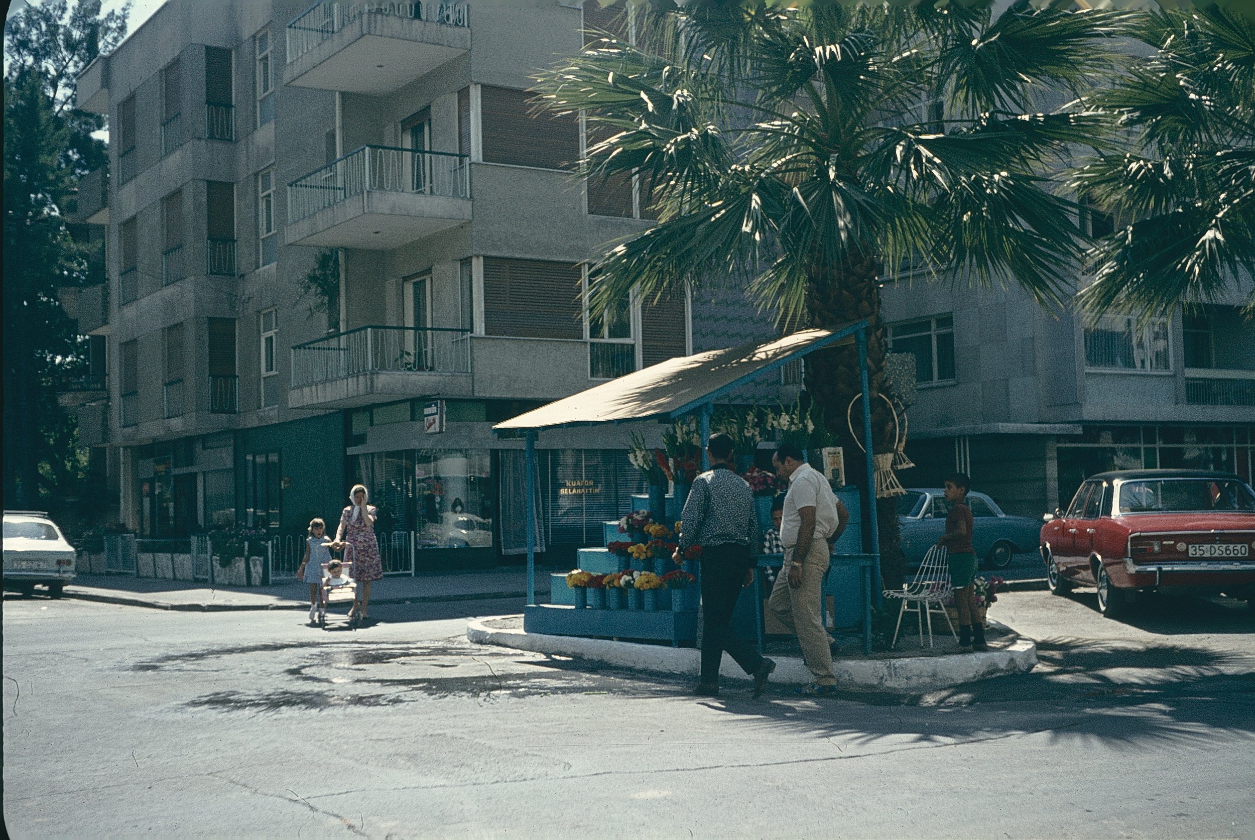 City street scene with a fruit stand and several people, including children, near a modern apartment building with balconies and large windows. A palm tree is prominently visible in the foreground.