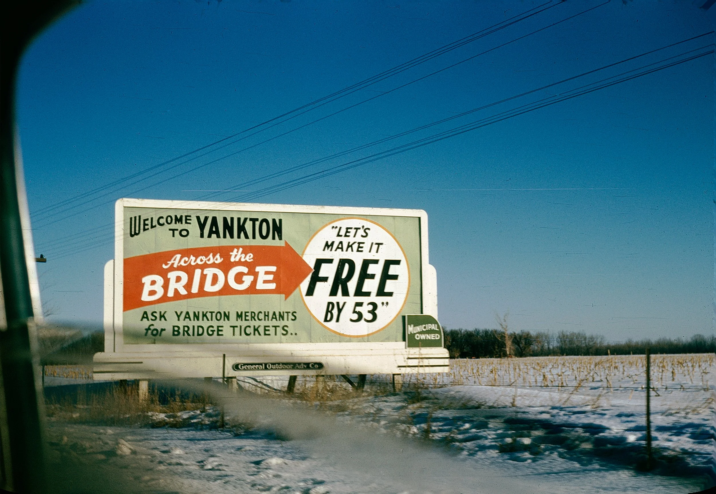 A billboard on the side of the road welcoming visitors to Yankton. It has a promotional message about making bridge tickets free by asking Yankton merchants. The sky is clear blue, and power lines are crossing above the billboard. Snow covers the gro