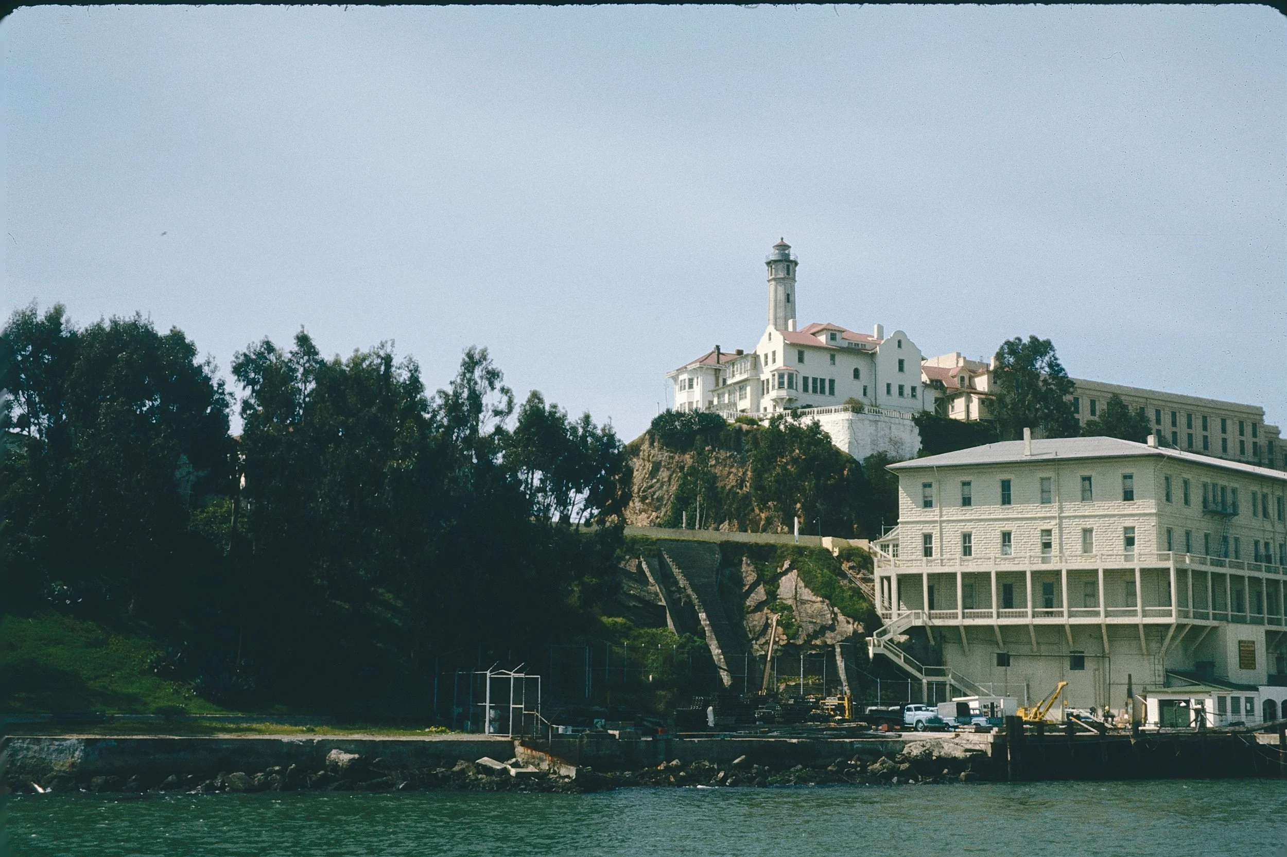A coastal area with a large white building featuring a tower and smaller structures, situated on a hillside among trees, with waterfront in the foreground.