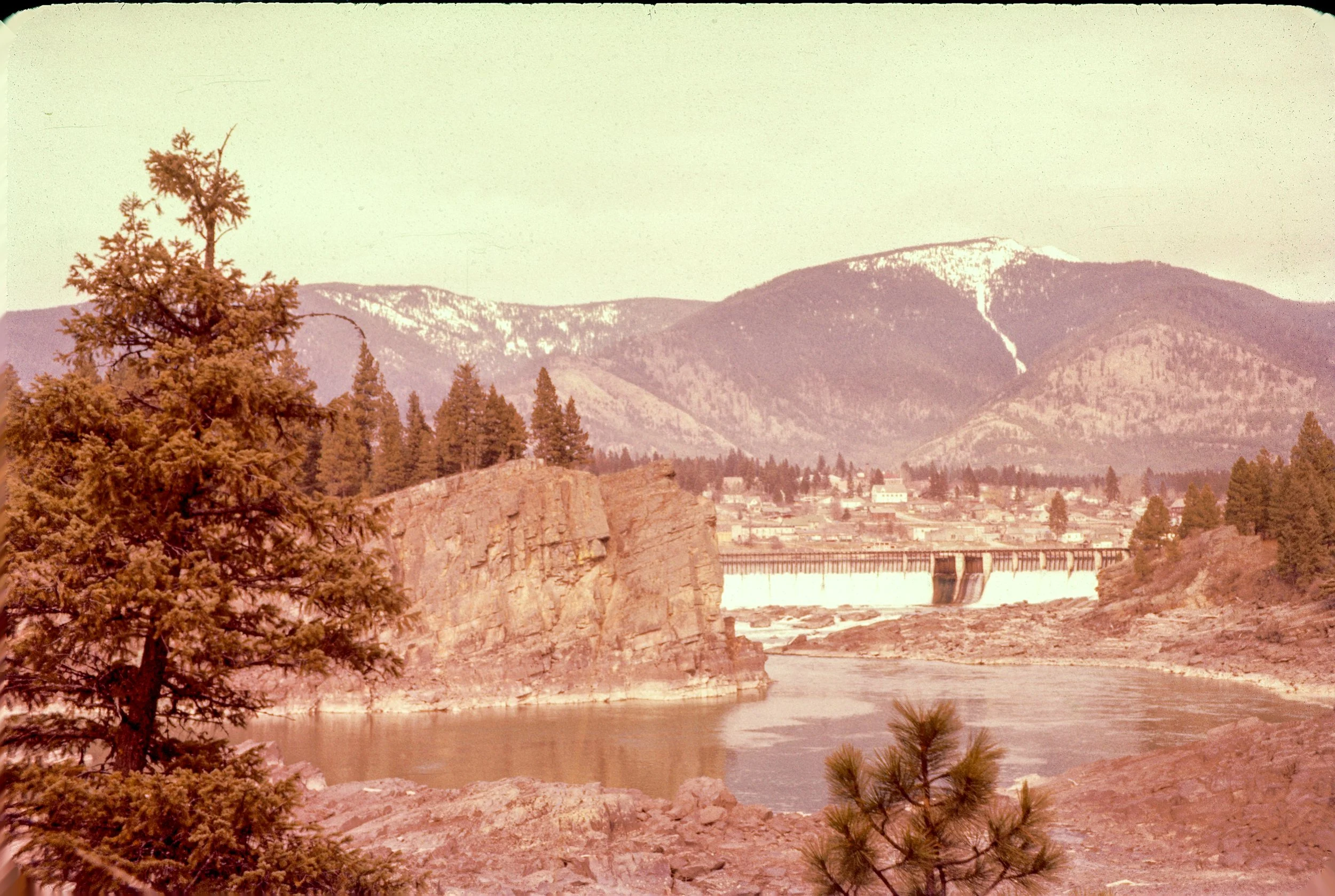 A river flowing through a rocky landscape with a dam and small town or village in the background. Mountainous terrain with somewhat snow-capped peaks in the distance. Several tall pine trees in the foreground.